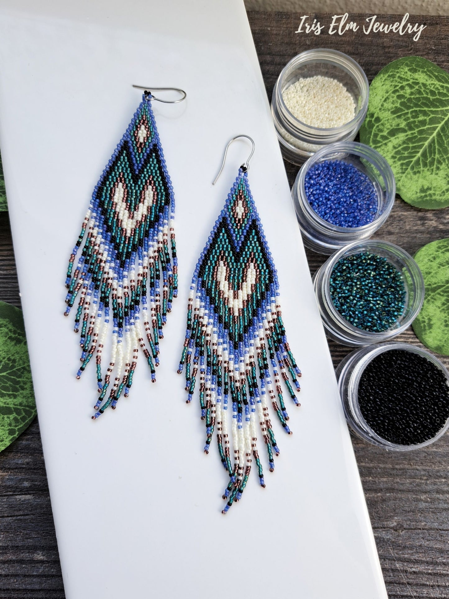 Beaded long purple, teal, black, and white seed bead heart fringe handmade earrings on a white surface with containers of beads and green leaves.