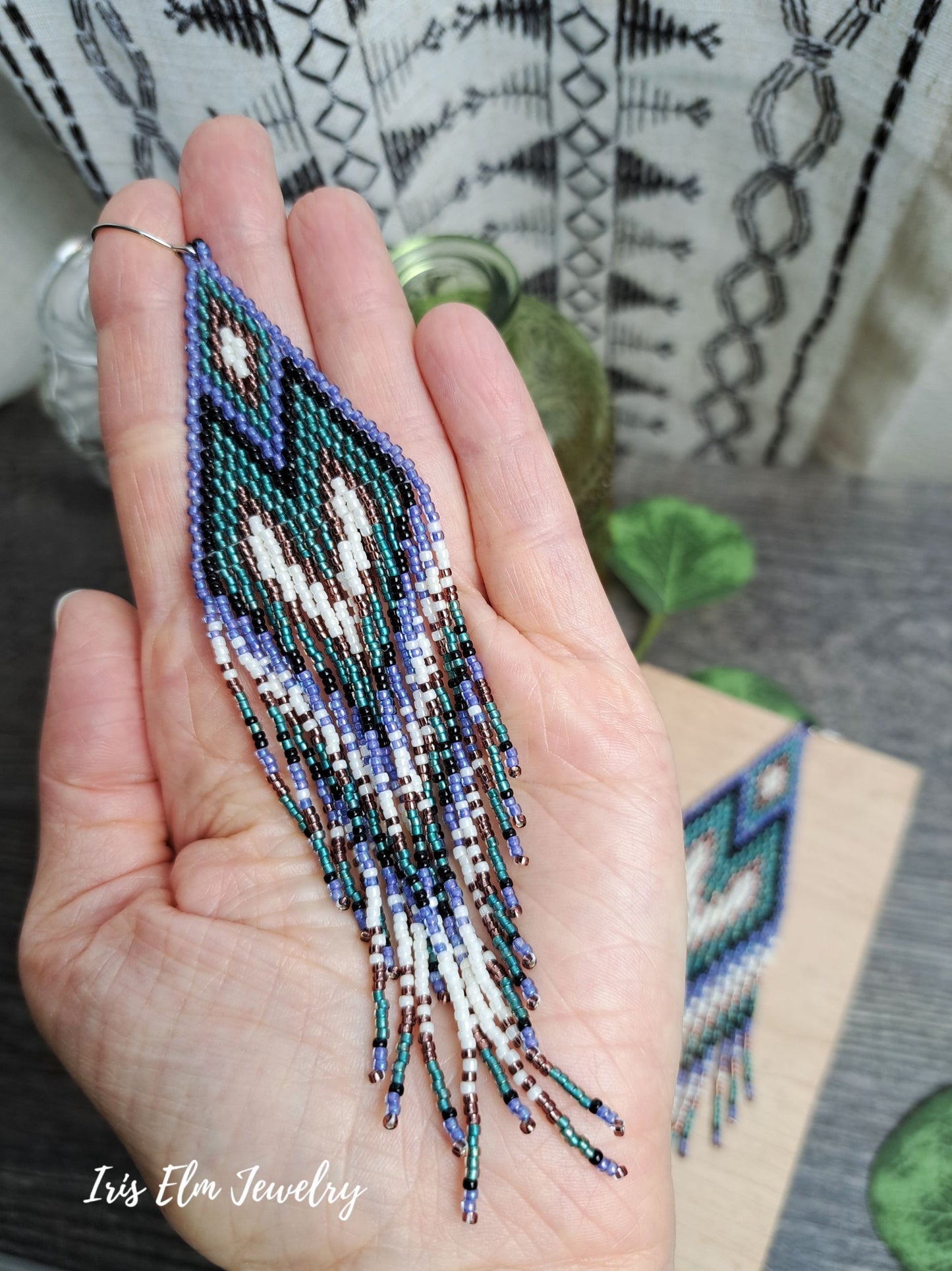 Hand holding a beadwork long purple, teal, black, and white seed bead heart fringe earrings with 'Iris Elm Jewelry' branding.