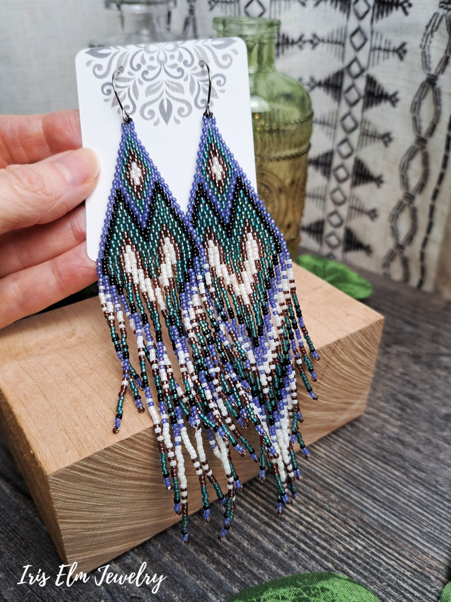 Beaded long purple, teal, black, and white seed bead heart fringe earrings on a display card held by a hand, with a blurred background