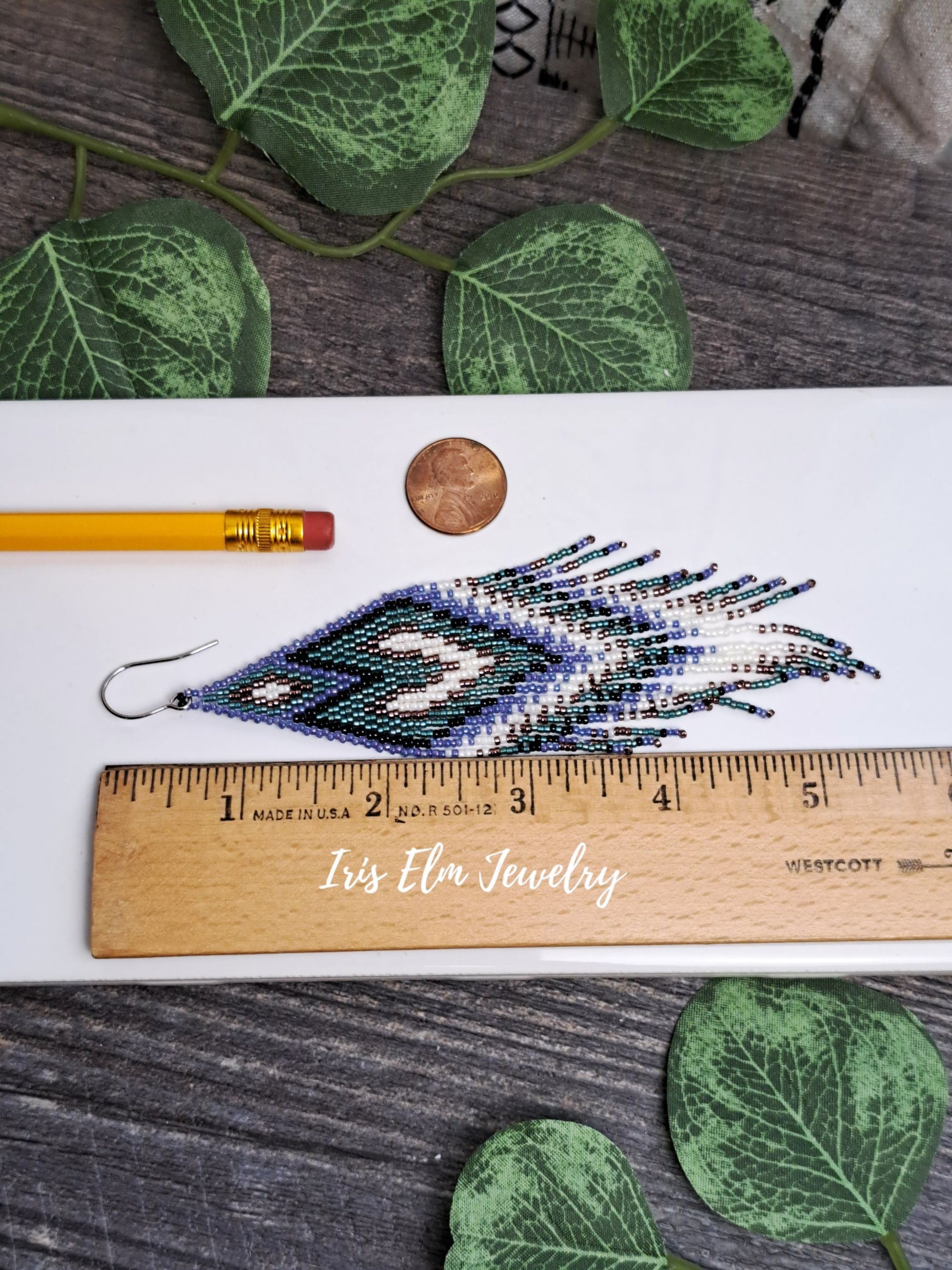 5.5 inch long seed bead fringe earring on a white surface with a ruler for scale, surrounded by green leaves.