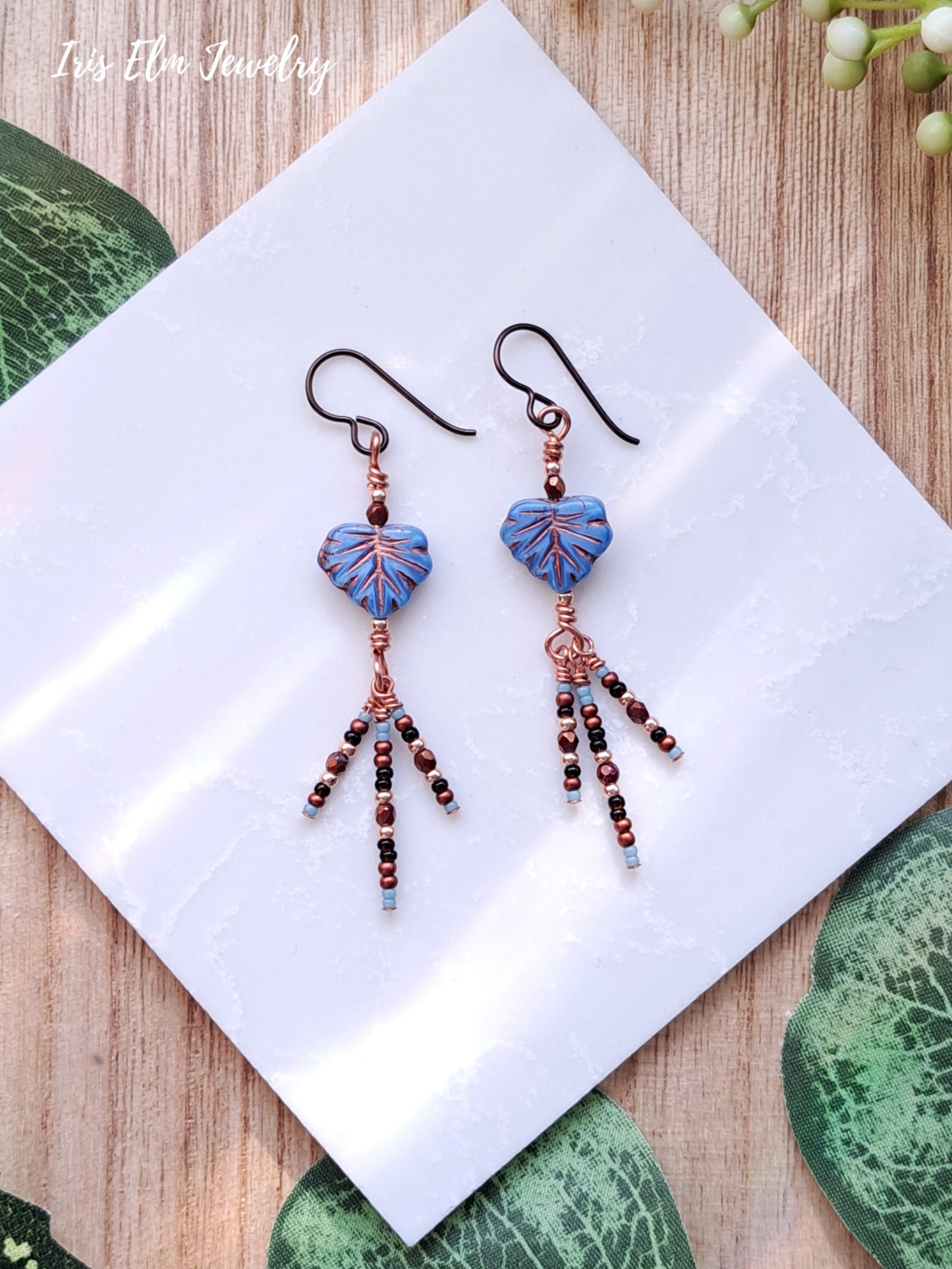 Pair of beaded blue leaf glass dangle earrings on a white napkin with green leaves in the background