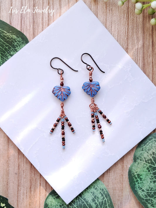 Pair of beaded blue leaf glass dangle earrings on a white napkin with green leaves in the background