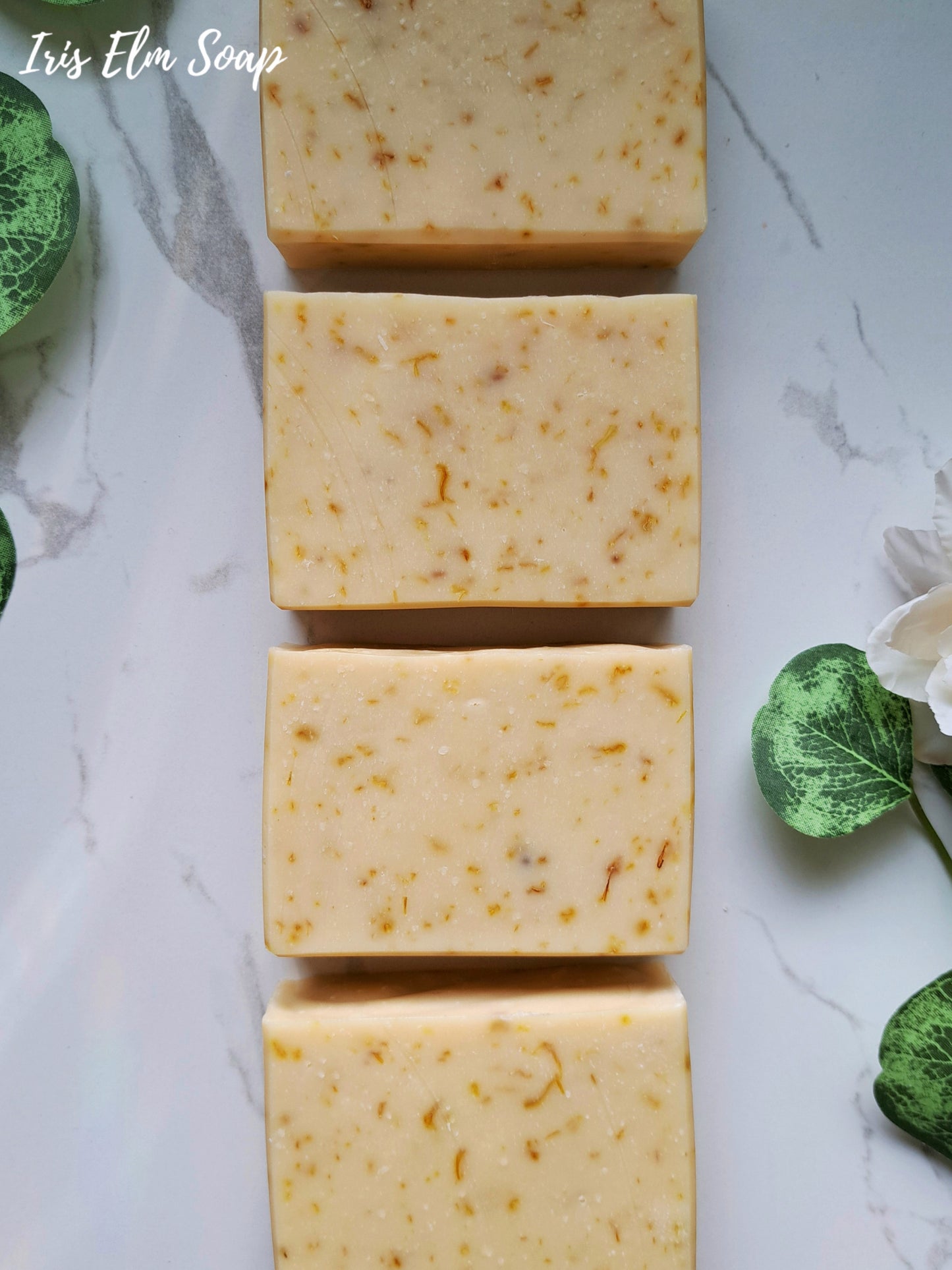 Four bars of orange calendula handmade soap on a marble surface with green leaves and flowers.