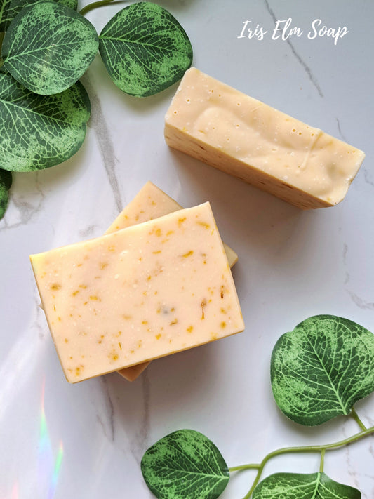 Two bars of yellow orange calendula soap on a marble surface with green leaves around, featuring 'Iris Elm Soap'.