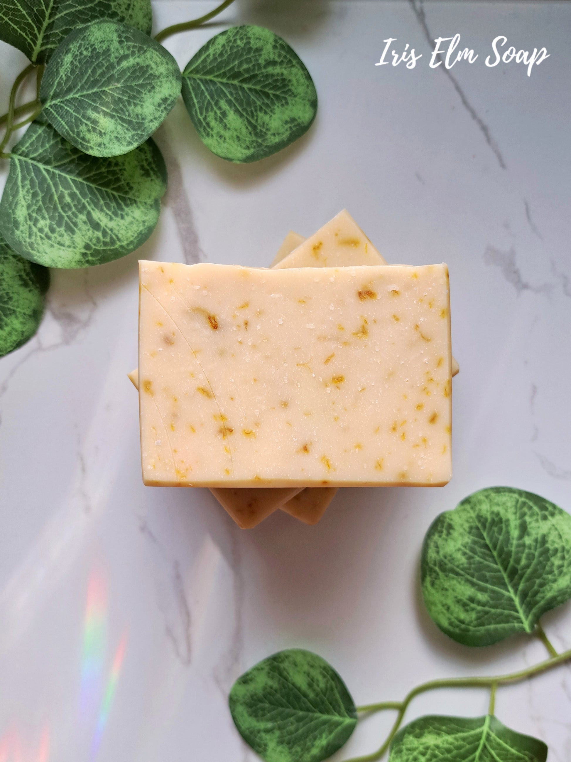 Two bars of orange calendula soap with visible calendula petals on a marble surface with green leaves.
