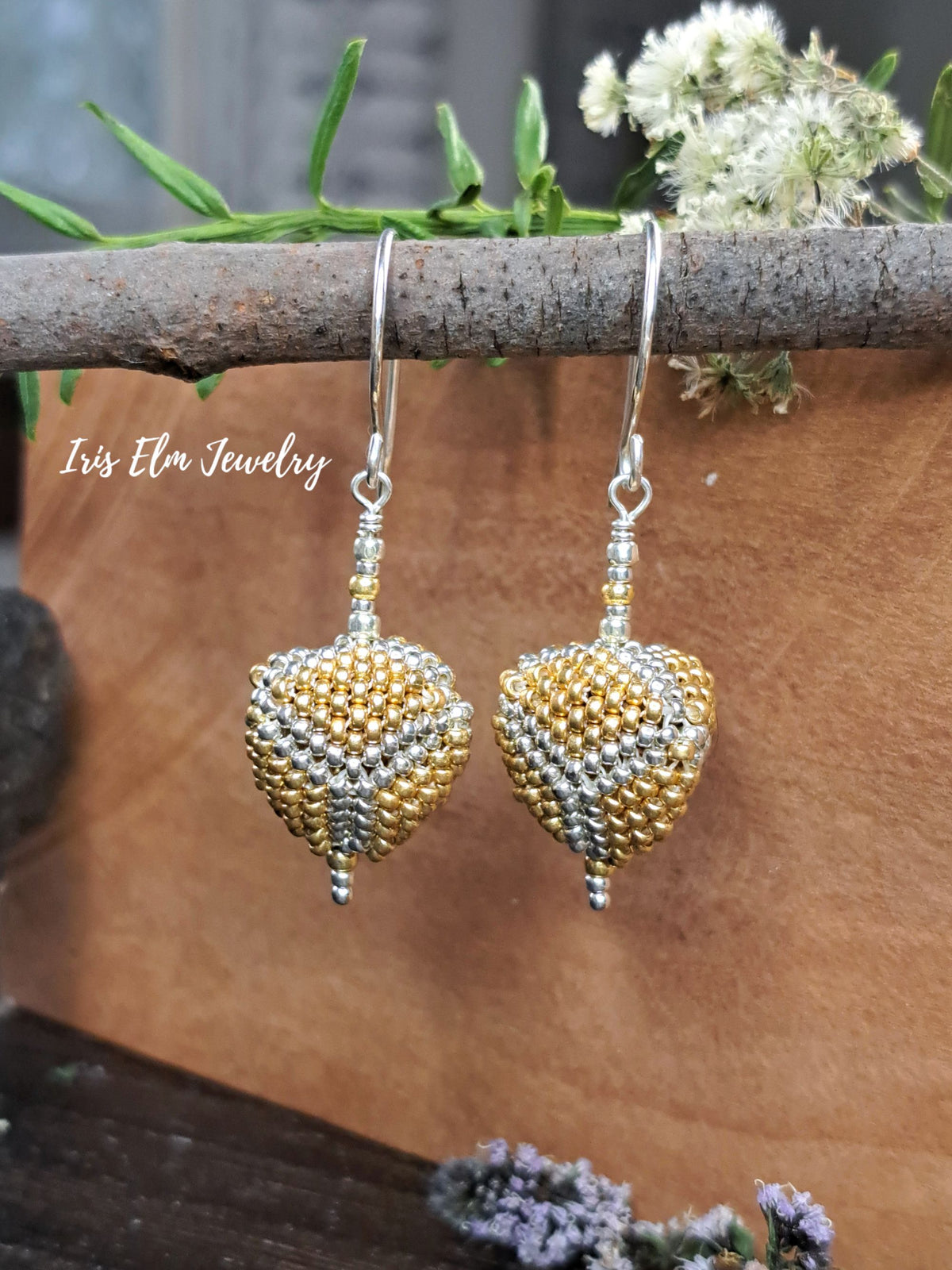 A pair of handmade beaded dangle earrings with gold and silver seed bead patterns, hanging from silver fishhook wires. The earrings are displayed against a brown paper background with a rustic branch, green leaves, and small flowers.