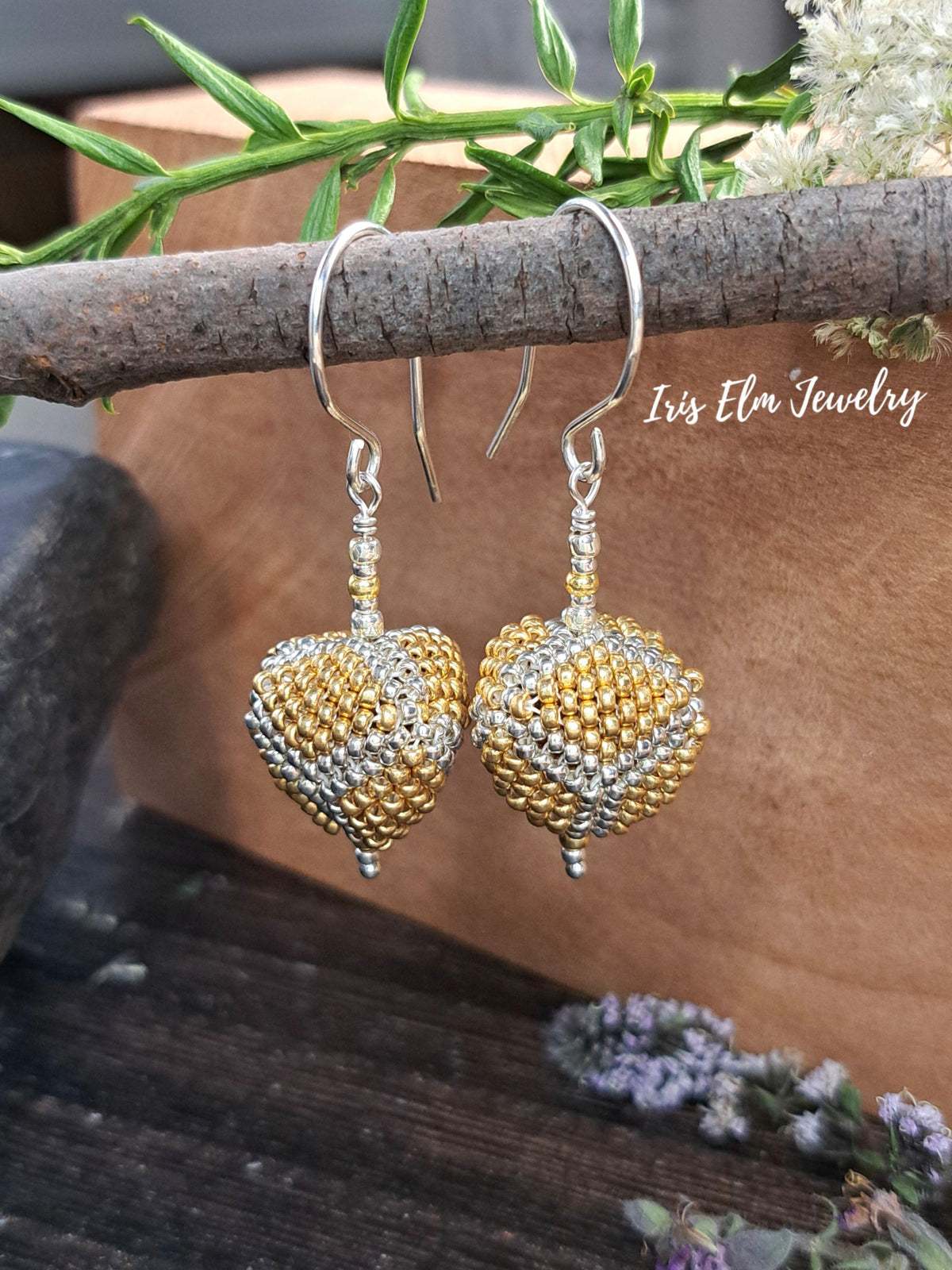 A pair of beaded dangle triangle earrings with gold and silver heart-shaped beads hanging from silver fishhook wires, displayed against a rustic brown background with green leaves and small purple flowers. The brand name 'Isis Elm Jewelry' is visible as a watermark.