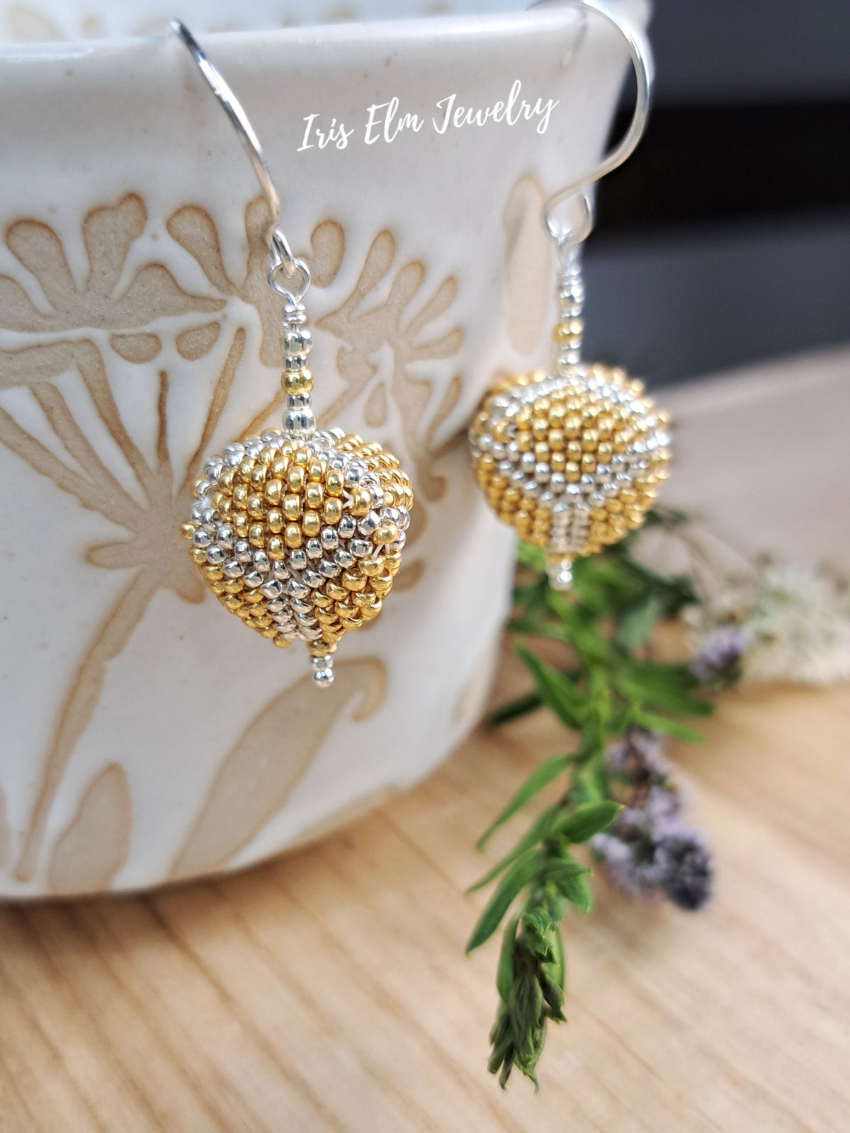 A pair of beaded dangle earrings with gold and silver seed bead patterns, hanging from silver fishhook wires. The earrings are displayed in front of a white ceramic mug with a beige floral pattern, with green sprigs on a wooden surface in the foreground.