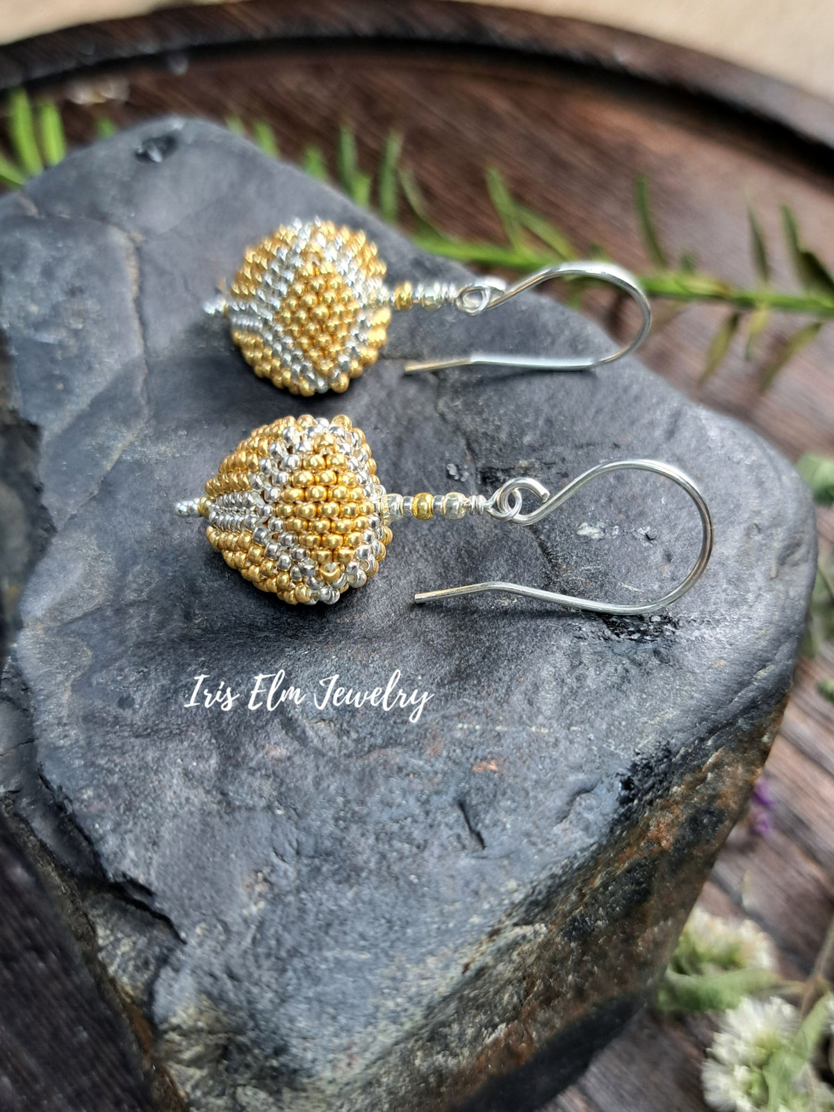A pair of handmade beaded dangle earrings with gold and silver seed beads on silver fishhook wires, displayed on a dark textured stone with green pine needles in the background. The brand name 'Iris Elm Jewelry' is watermarked on the stone.