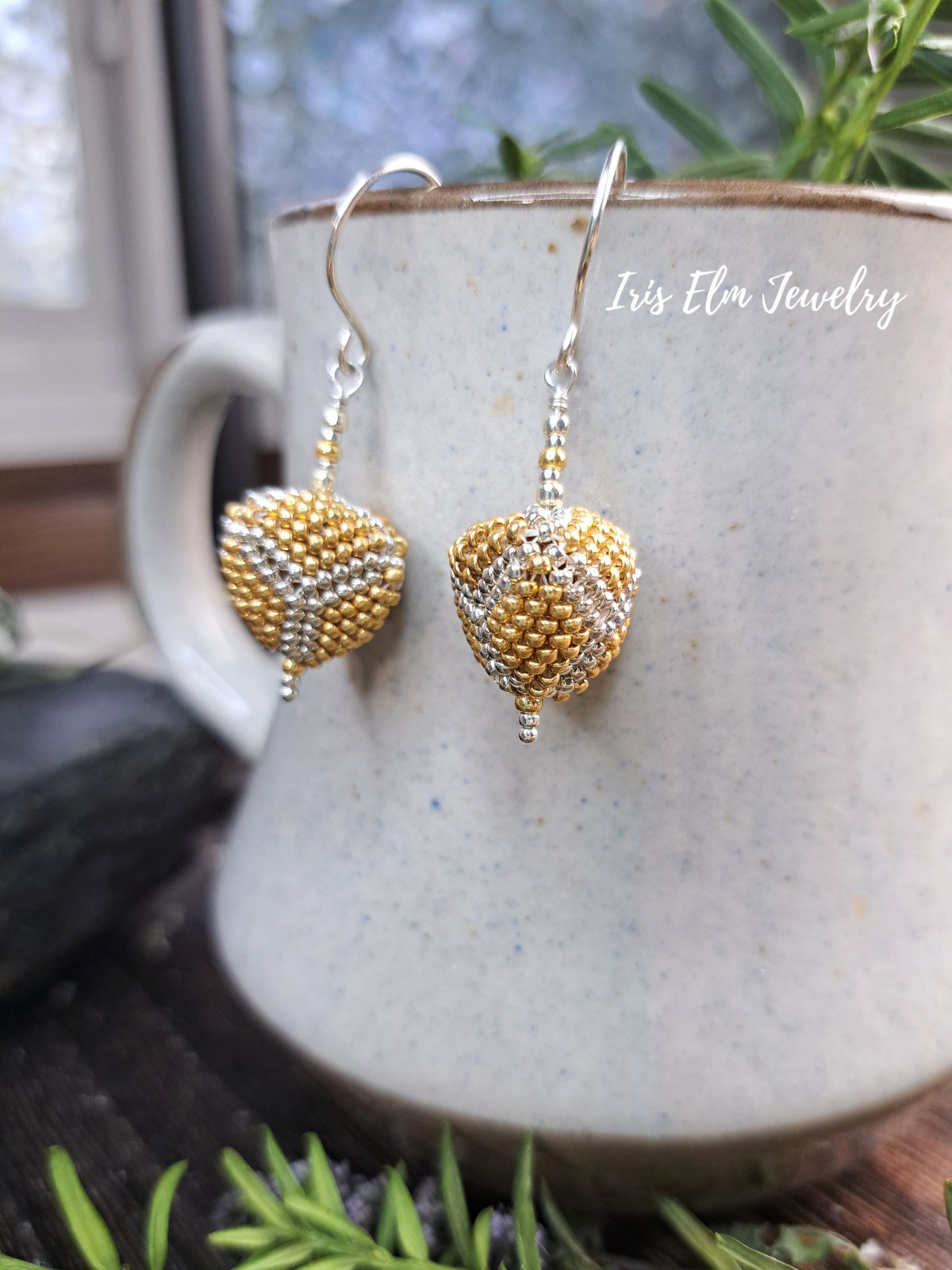 A pair of handmade beaded dangle earrings with gold and silver seed beads forming heart-like shapes, hanging from silver fishhook wires. The earrings are displayed on a speckled ceramic mug with green foliage in the background.