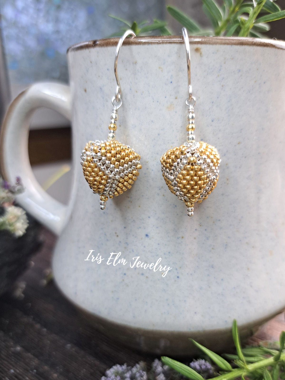 A pair of handmade beaded heart-shaped drop earrings in gold and silver, hanging from silver fishhook wires. They are displayed draped over a speckled ceramic mug with the brand name 'Iris Elm Jewelry' printed on it, surrounded by green sprigs and small flowers.