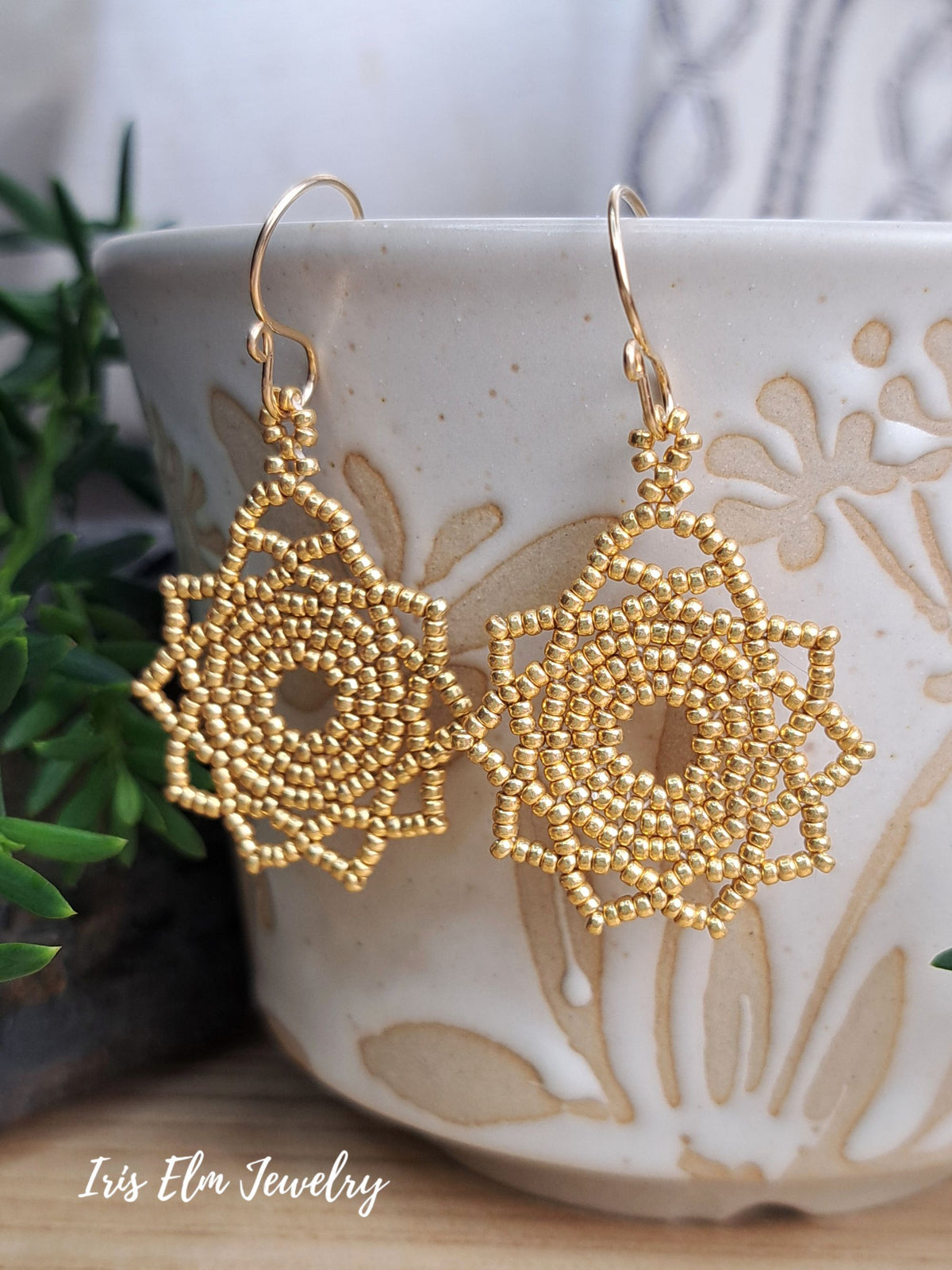 A pair of gold beaded dangle earrings with a geometric, lace-like starburst mandala design, hanging from gold fishhook wires. They are displayed resting on a textured beige ceramic cup with embossed floral patterns, with green foliage in the background.