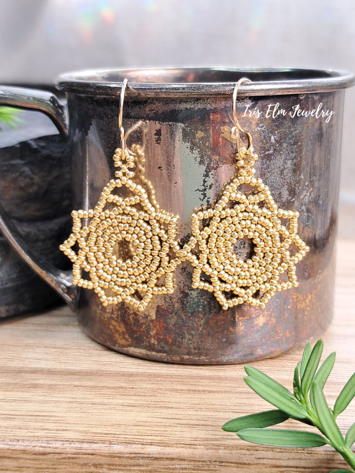 A pair of handmade bohemian beaded dangle earrings in a gold tone, hanging from a rustic, distressed metal mug on a wooden surface with a green sprig in the foreground.