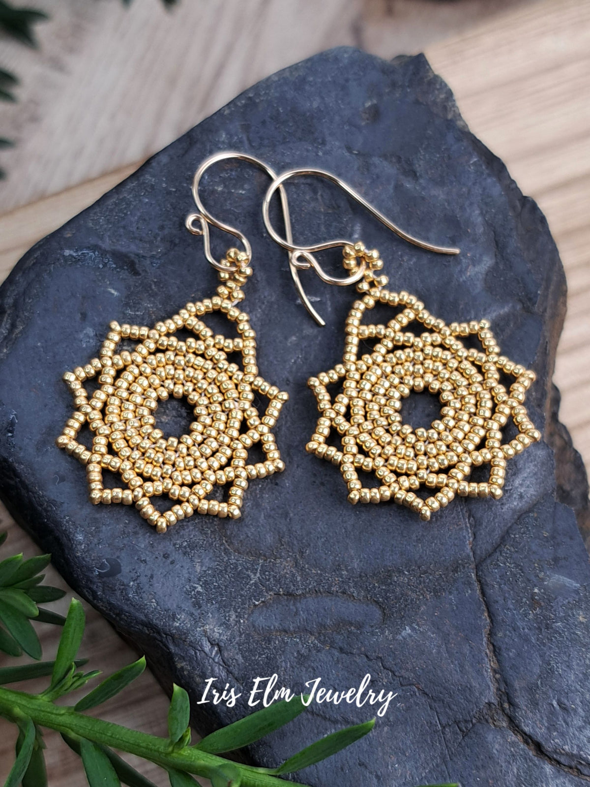 A pair of handmade gold beaded dangle earrings with a geometric spiral pattern, displayed on a dark slate stone with green leaves and a wooden surface in the background. The brand name 'Iris Elm Jewelry' is watermarked at the bottom.