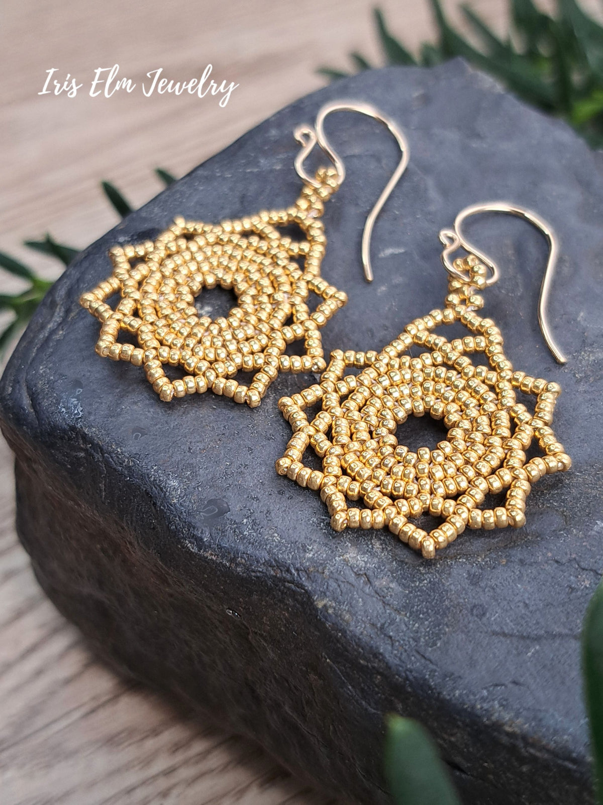 A pair of gold beaded dangle earrings with a geometric mandala-like pattern, resting on a dark slate block with green foliage in the background. The brand name 'Iris Elm Jewelry' is visible in the top left corner.