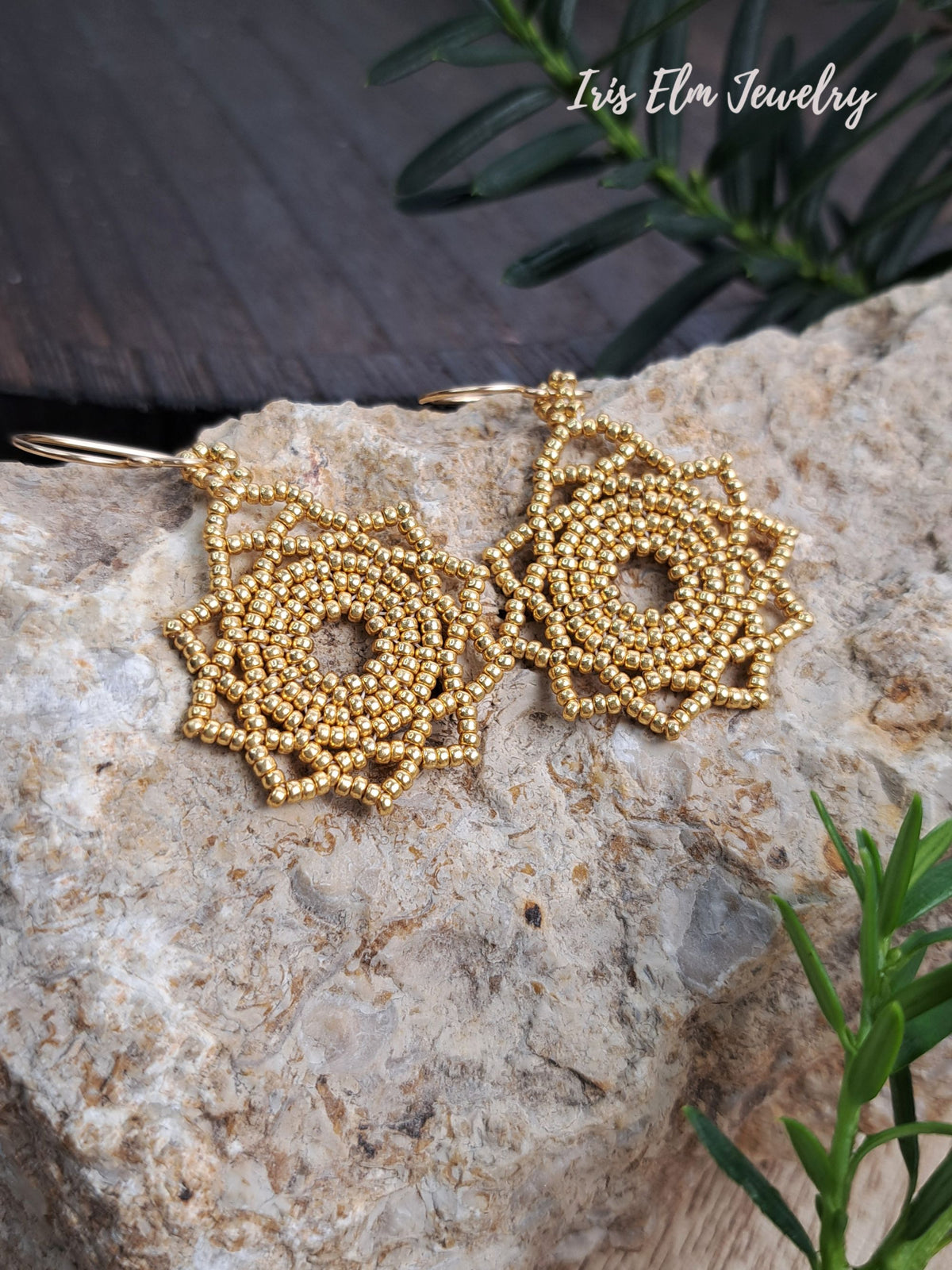 A pair of gold beaded hoop earrings with intricate spiral patterns, displayed on a textured stone surface with green foliage in the background. The brand name 'Iris Elm Jewelry' is visible in the top right corner.