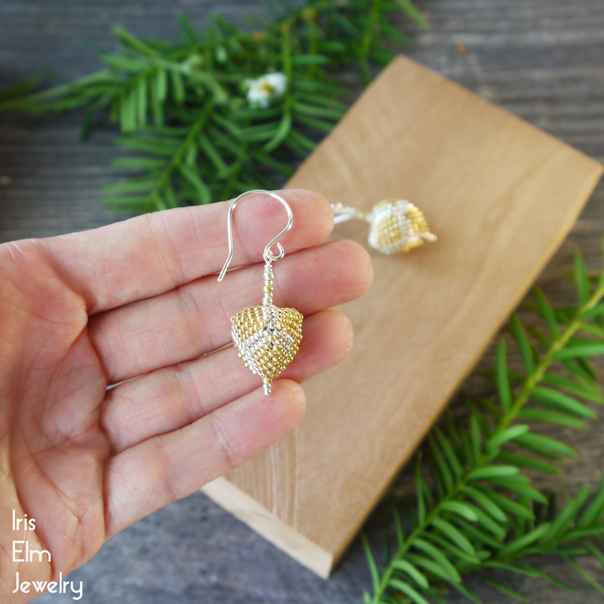 Gold and silver 3d triangle seed bead earrings held in a hand with a wooden block and greenery in the background.
