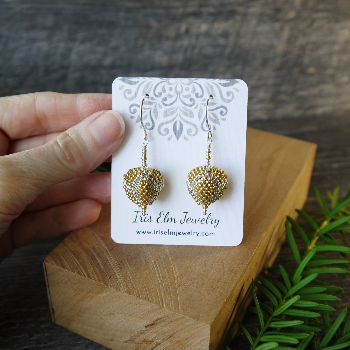 Gold triangle-shaped earrings on a 'Iris Elm Jewelry' card held by a hand with a wooden block and plant in the background.