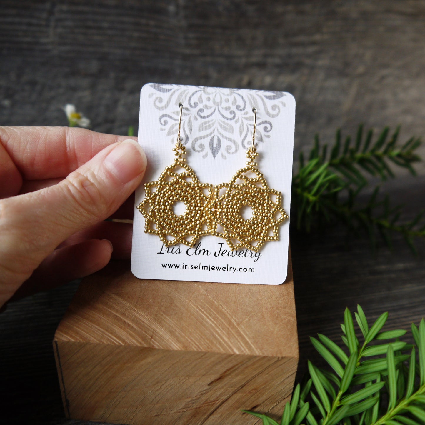 Gold woven mandala earrings on a 'Iris Elm Jewelry' branded card held by a hand with a wooden block and green leaves in the background