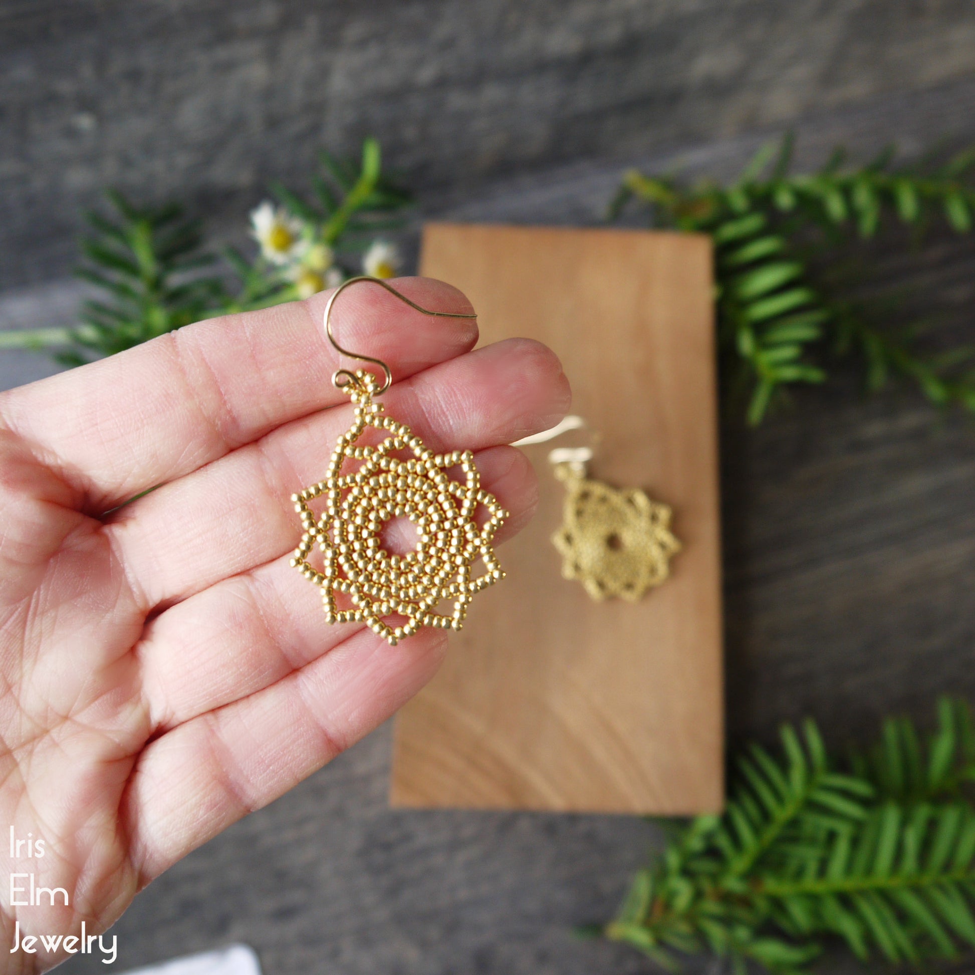 Gold beaded circular mandala earrings held in a hand with a wooden box and greenery in the background.