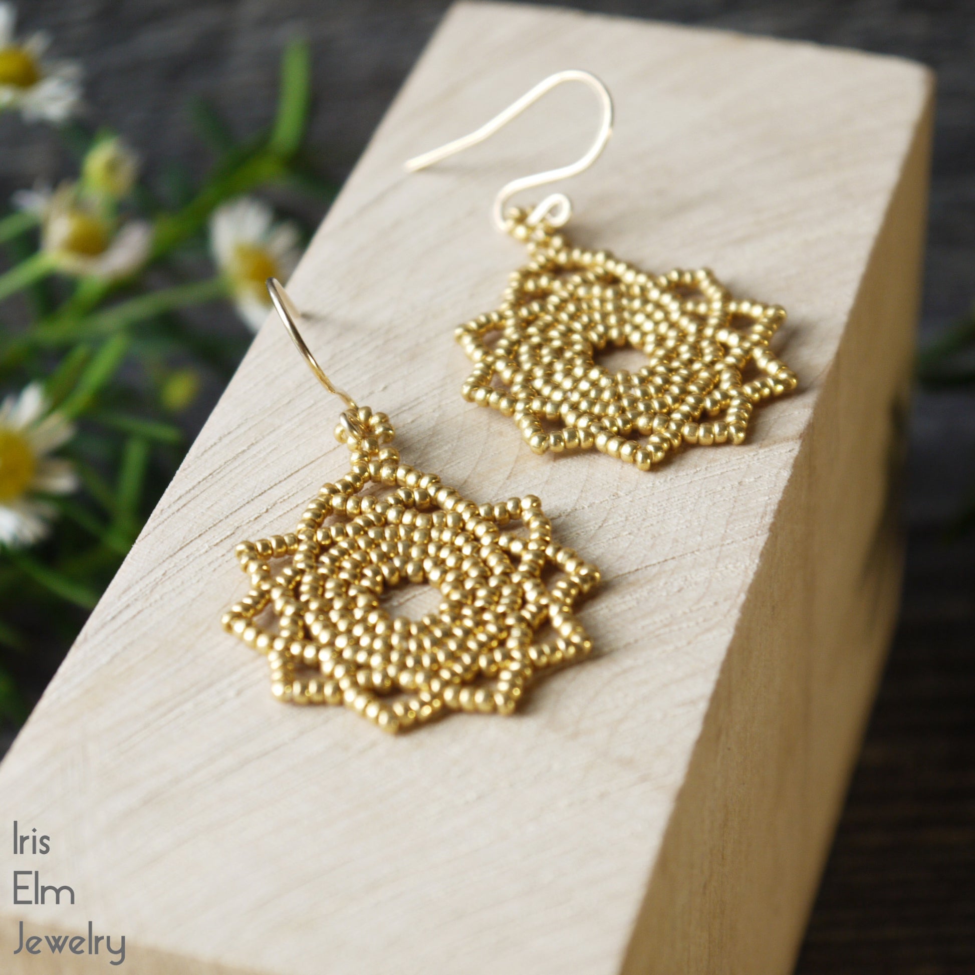 handcrafted Gold beaded earrings on a wooden block with blurred flowers in the background