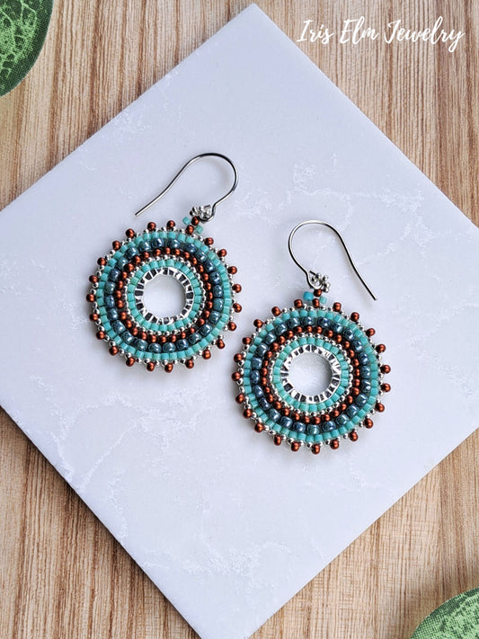 A pair of handmade beaded drop earrings with turquoise, brown, and white circular patterns on silver fishhook wires, displayed on a white square card over a wooden surface.