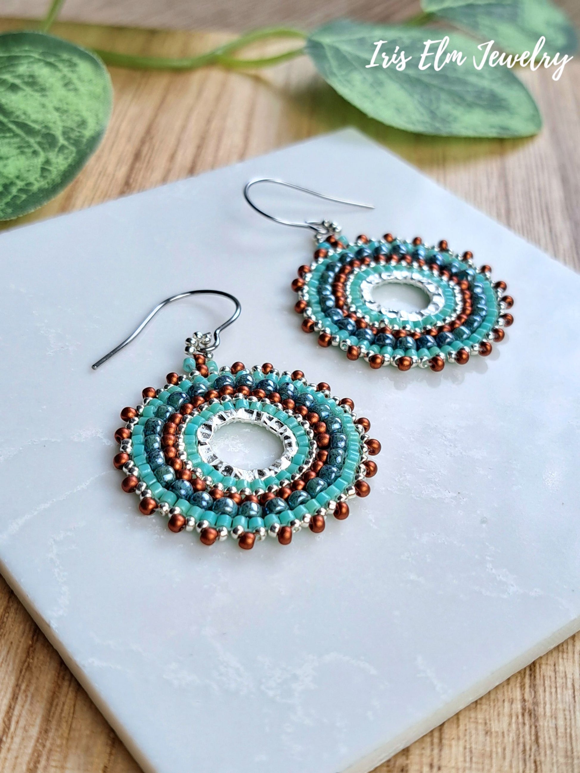 A pair of handmade beaded dangle earrings with turquoise, brown, and silver beads, displayed on a white marble tile with green leaves in the background. The brand name 'Iris Elm Jewelry' is visible in the top right corner.