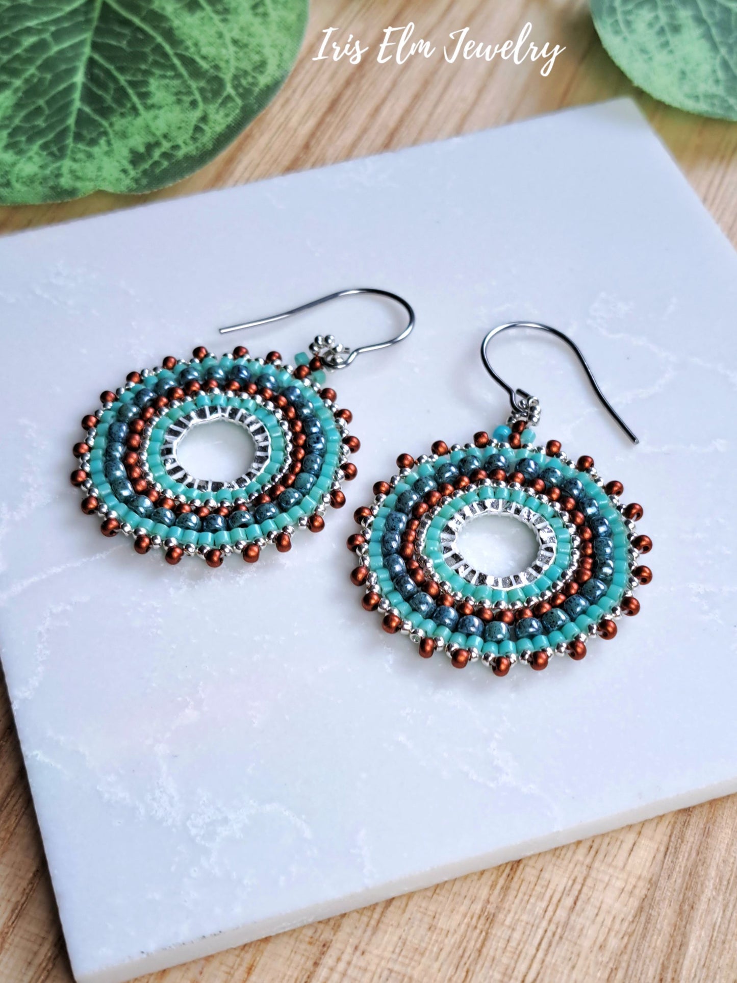 A pair of handmade beaded dangle earrings with turquoise, brown, and silver beads, displayed on a white marble tile with green leaves in the background. The text 'Iris Elm Jewelry' is visible in the top right corner.