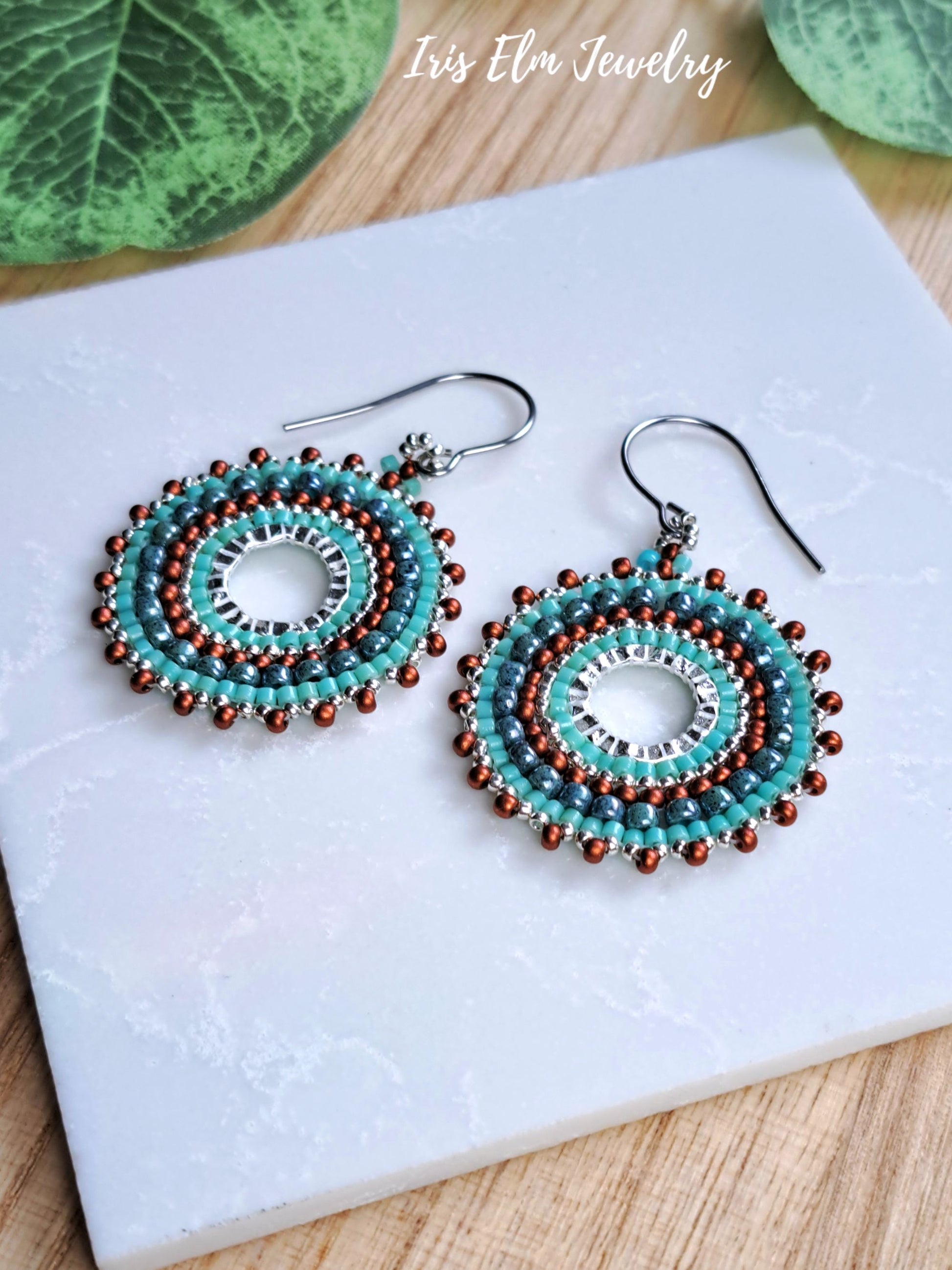 A pair of handmade beaded dangle earrings with turquoise, brown, and silver beads, displayed on a white marble tile with green leaves in the background. The text 'Iris Elm Jewelry' is visible in the top right corner.