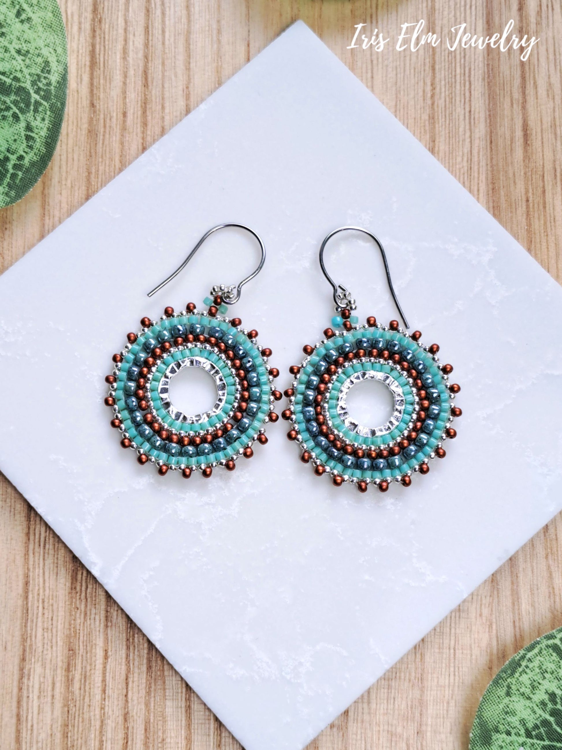 A pair of handmade beaded hoop earrings with turquoise, white, and brown seed beads on silver fishhook wires, displayed on a white textured square card over a wooden surface.