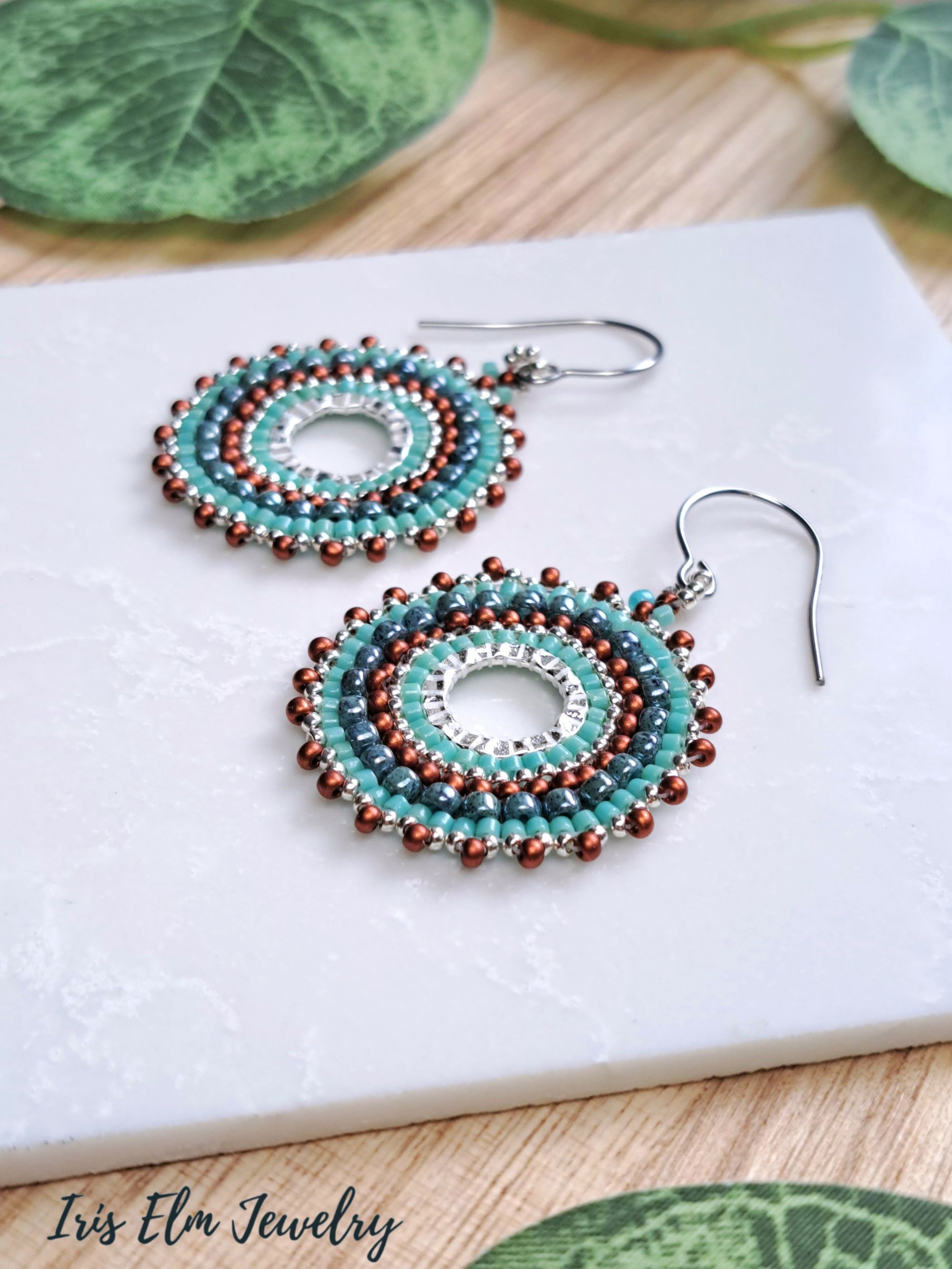 A pair of handmade beaded dangle earrings with turquoise, brown, and silver beads, displayed on a white textured tile with green leaves in the background. The brand name 'Iris Elm Jewelry' is visible in the bottom left corner.