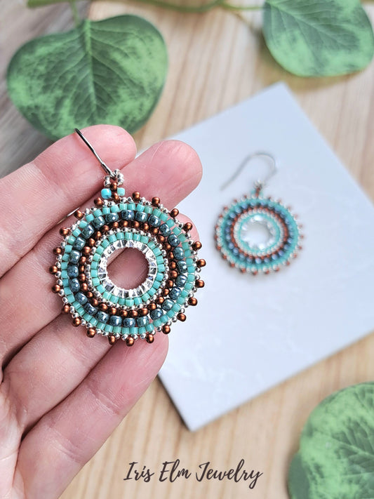 A hand holds a pair of handmade beaded earrings with turquoise, white, and copper-colored beads, with a matching pair resting on a white card in the background. Green leaves and a wooden surface frame the scene.