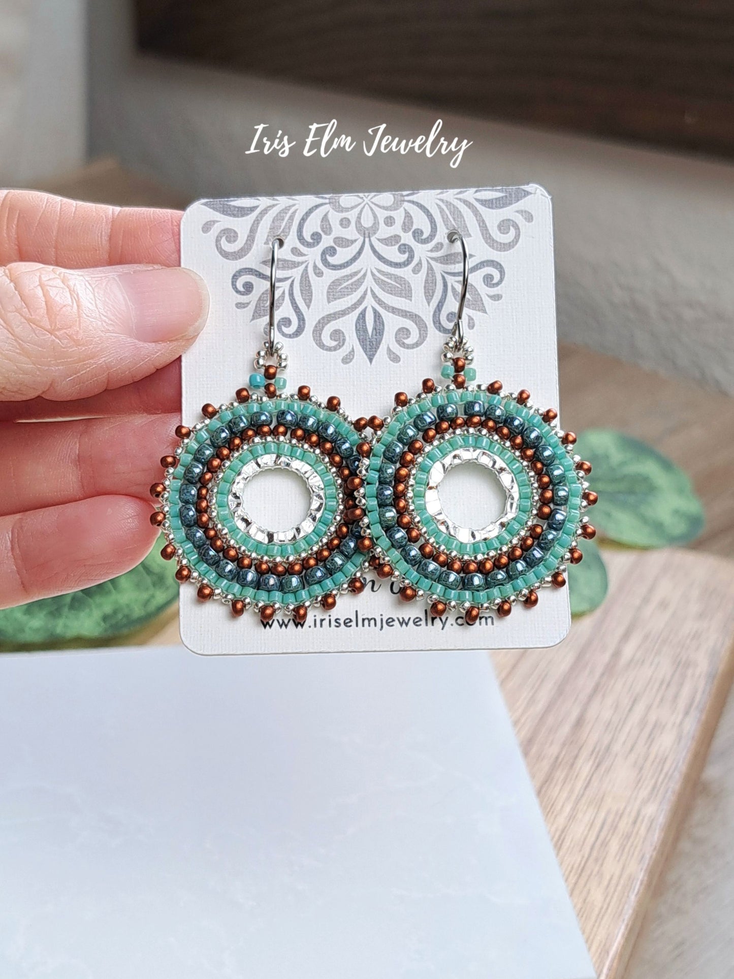 A pair of handmade beaded dangle earrings held by a hand on a white display card. The earrings feature turquoise, brown, and white seed beads in a circular pattern with silver-toned hooks.