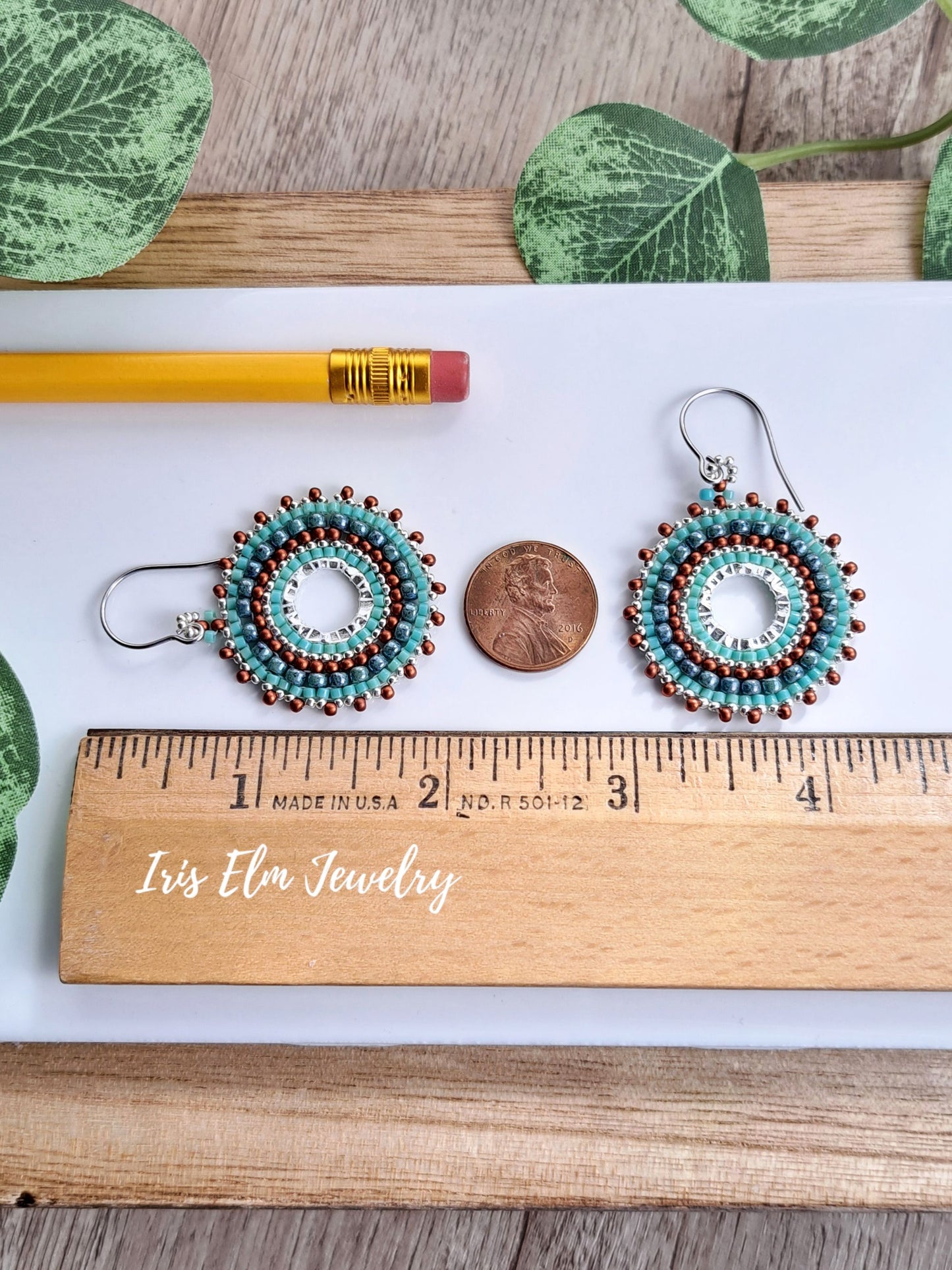 A pair of handmade beaded 2 inch long earrings with turquoise, white, and brown beads, displayed on a white card with a wooden ruler, a pencil, a penny, and green leaves. The brand name 'Iris Elm Jewelry' is printed on the ruler.