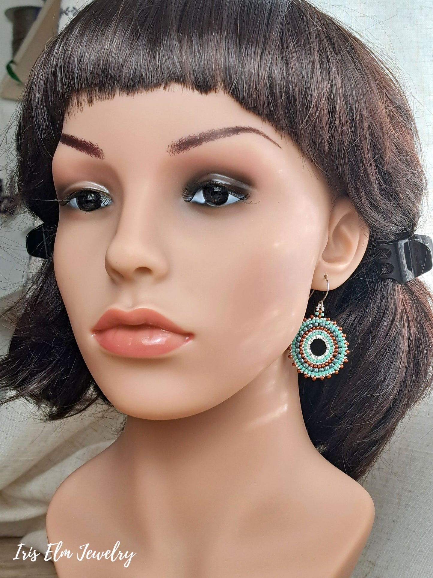 A close-up of a mannequin bust wearing a dangling circular earring with turquoise, white, and copper concentric details. The mannequin has dark brown hair with bangs.