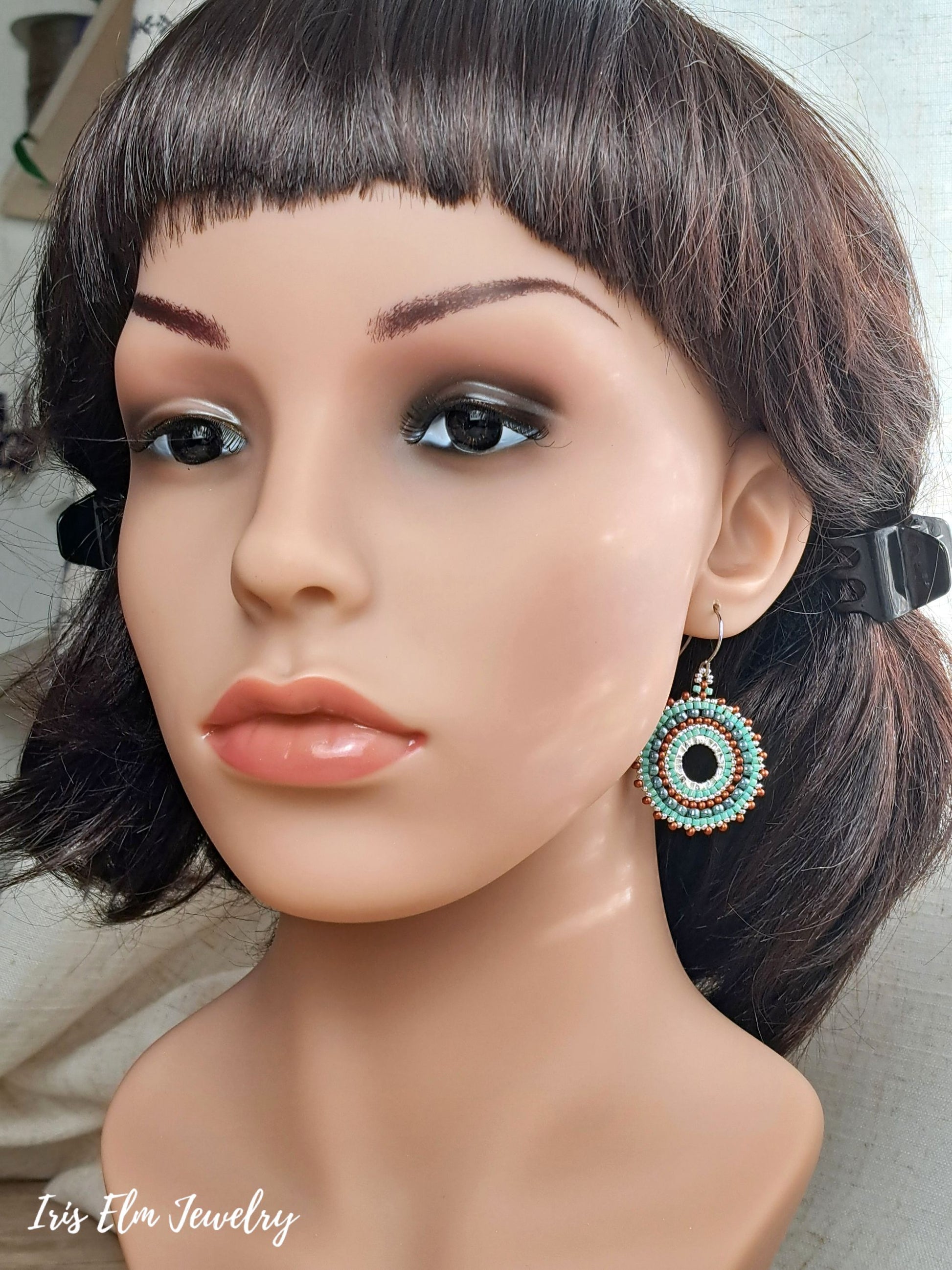 A close-up of a mannequin bust wearing a dangling circular earring with turquoise, white, and copper concentric details. The mannequin has dark brown hair with bangs.