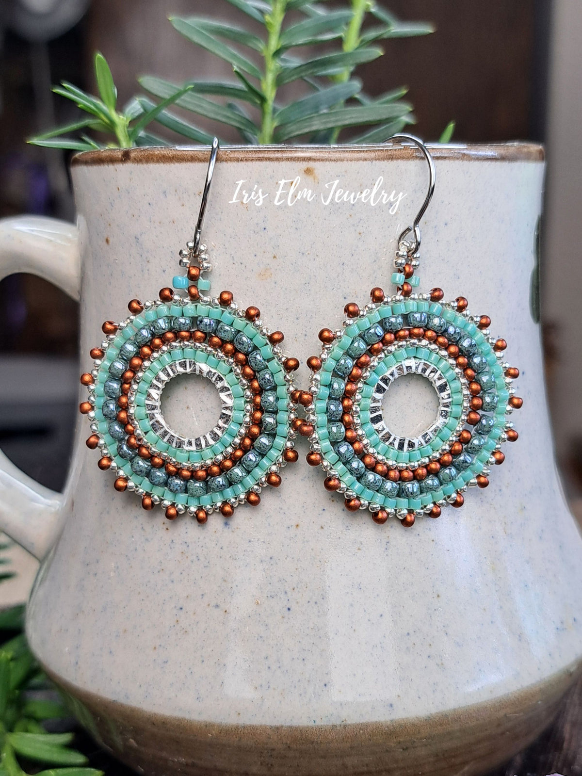 A pair of handmade beaded dangle earrings with turquoise, silver, and copper-colored beads, hanging from silver fishhook wires. The earrings are displayed on the rim of a speckled ceramic mug with green foliage in the background.