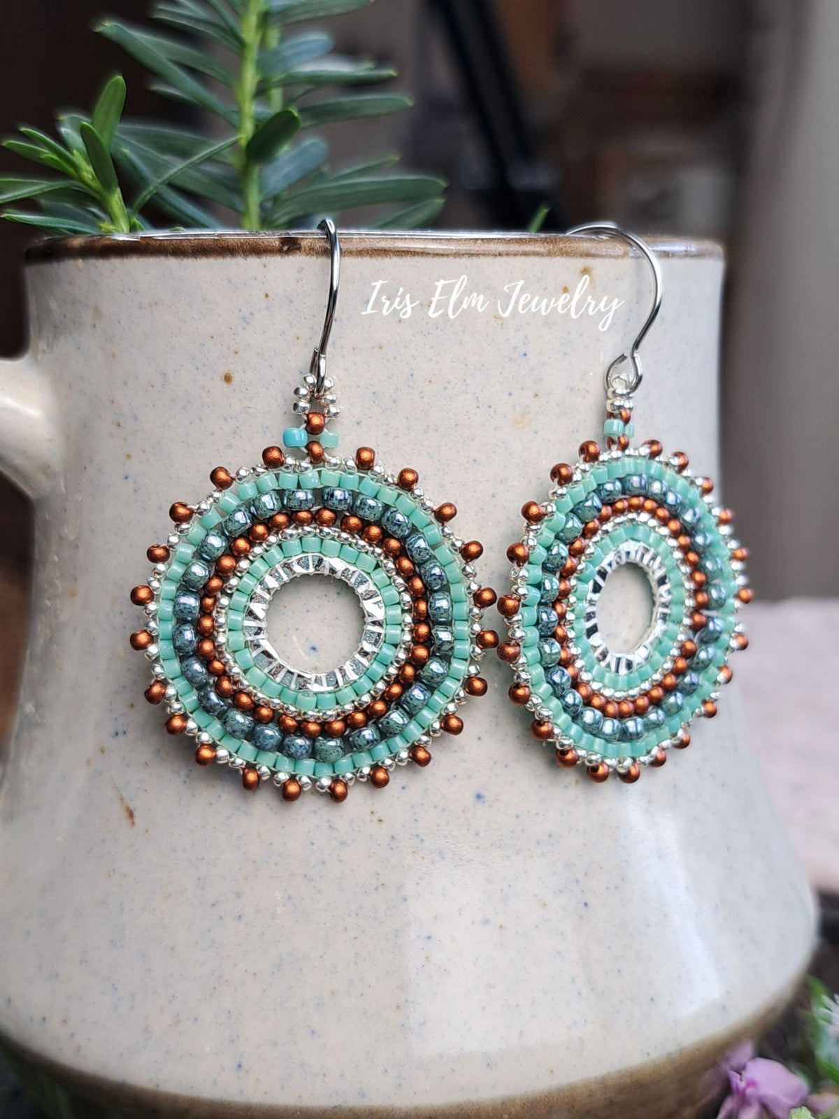 A pair of handmade circular beaded dangle earrings with turquoise, silver, and copper-colored beads, hanging from silver fishhook wires. They are displayed on a speckled beige ceramic mug with a green plant in the background.