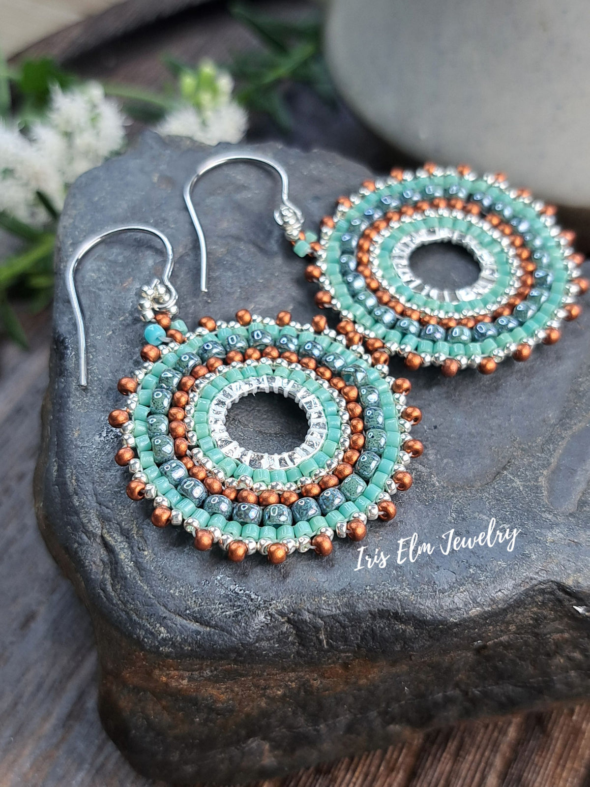 A pair of handmade beaded dangle earrings displayed on a dark stone surface. The earrings feature concentric circular patterns with turquoise, silver, and copper-colored beads, suspended from silver fishhook wires.