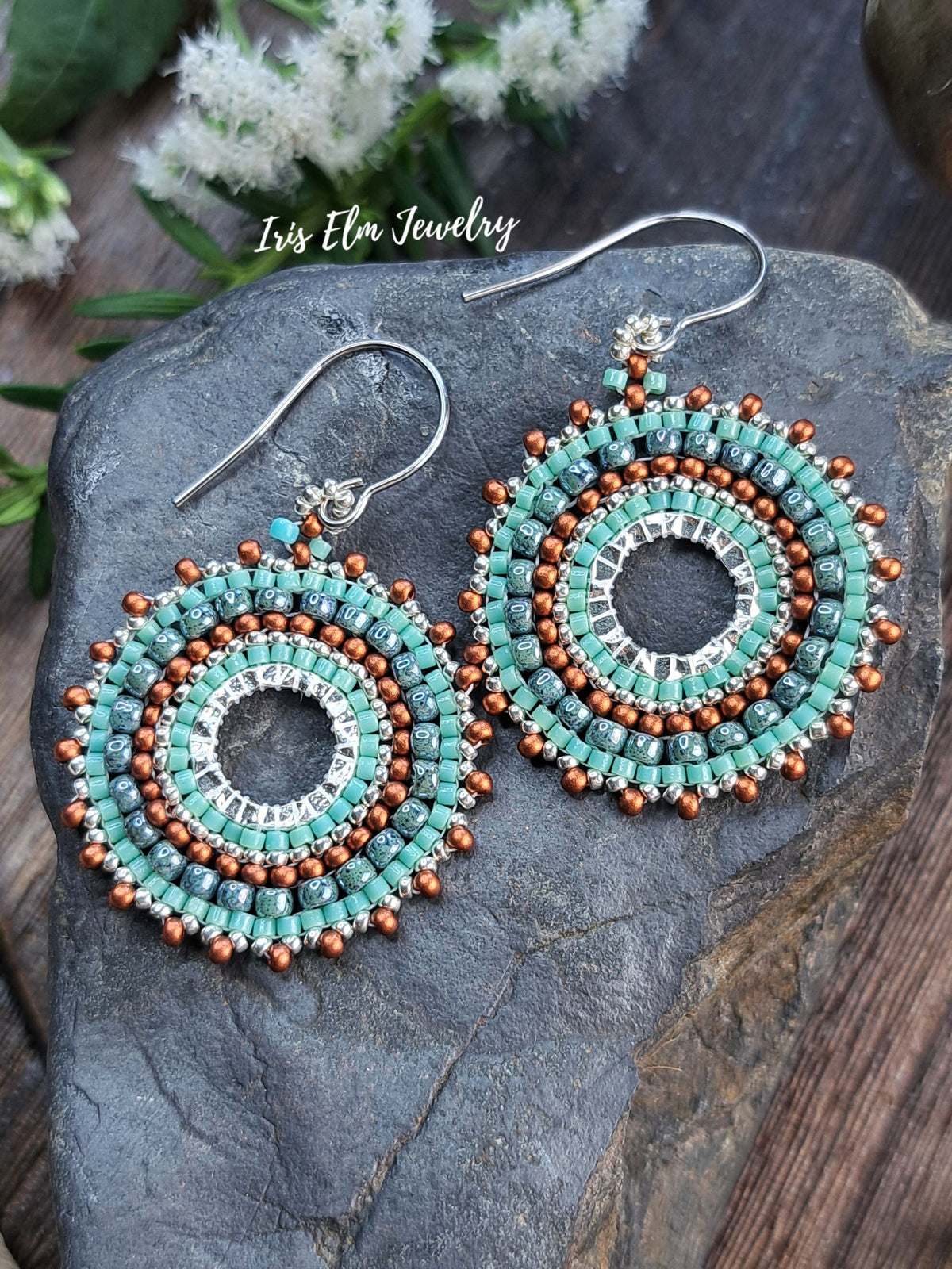 A pair of handmade beaded dangle earrings by Isis Elm Jewelry, featuring turquoise, brown, and silver beads in a circular spiral design, displayed on a dark gray stone.
