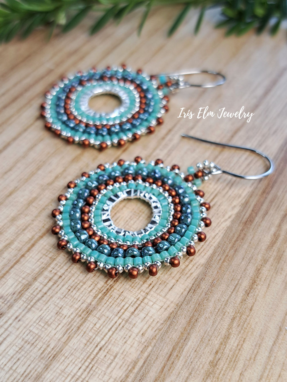 A pair of handmade beaded dangle earrings with turquoise, brown, and silver beads, displayed on a wooden surface with a green pine branch in the background. The brand name 'Iris Elm Jewelry' is visible as a watermark.