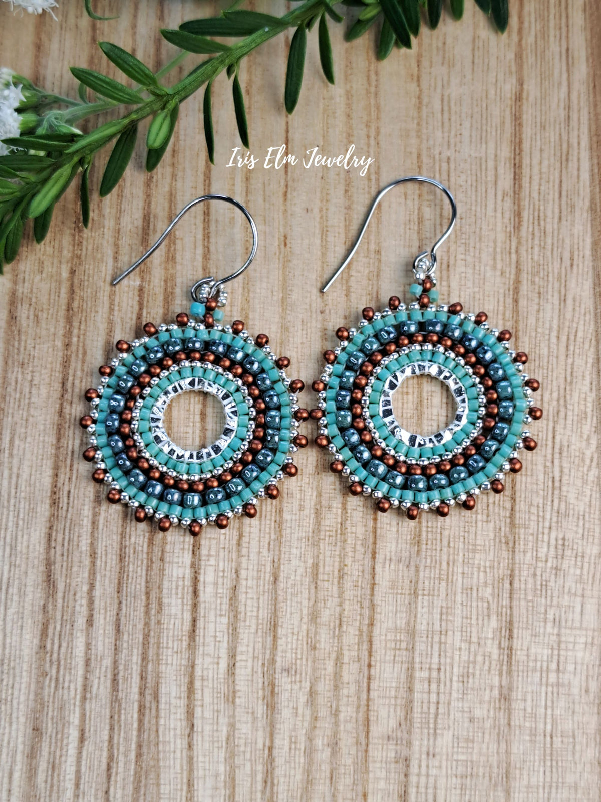 A pair of handmade beaded dangle earrings by Iris Elm Jewelry, featuring turquoise, brown, and silver seed beads in concentric circular patterns with silver fishhook wires, displayed on a light wood background with green leaves.