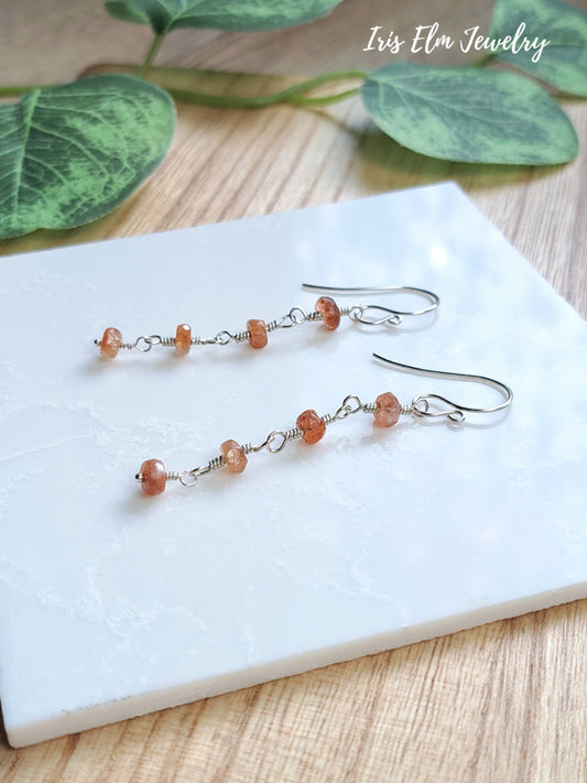 A pair of dangling earrings with small peach-orange sunstone beads on sterling silver hooks, displayed on a white rectangular tray over a light wood surface with green leaves in the background. The text 'Iris Elm Jewelry' appears in the top right.