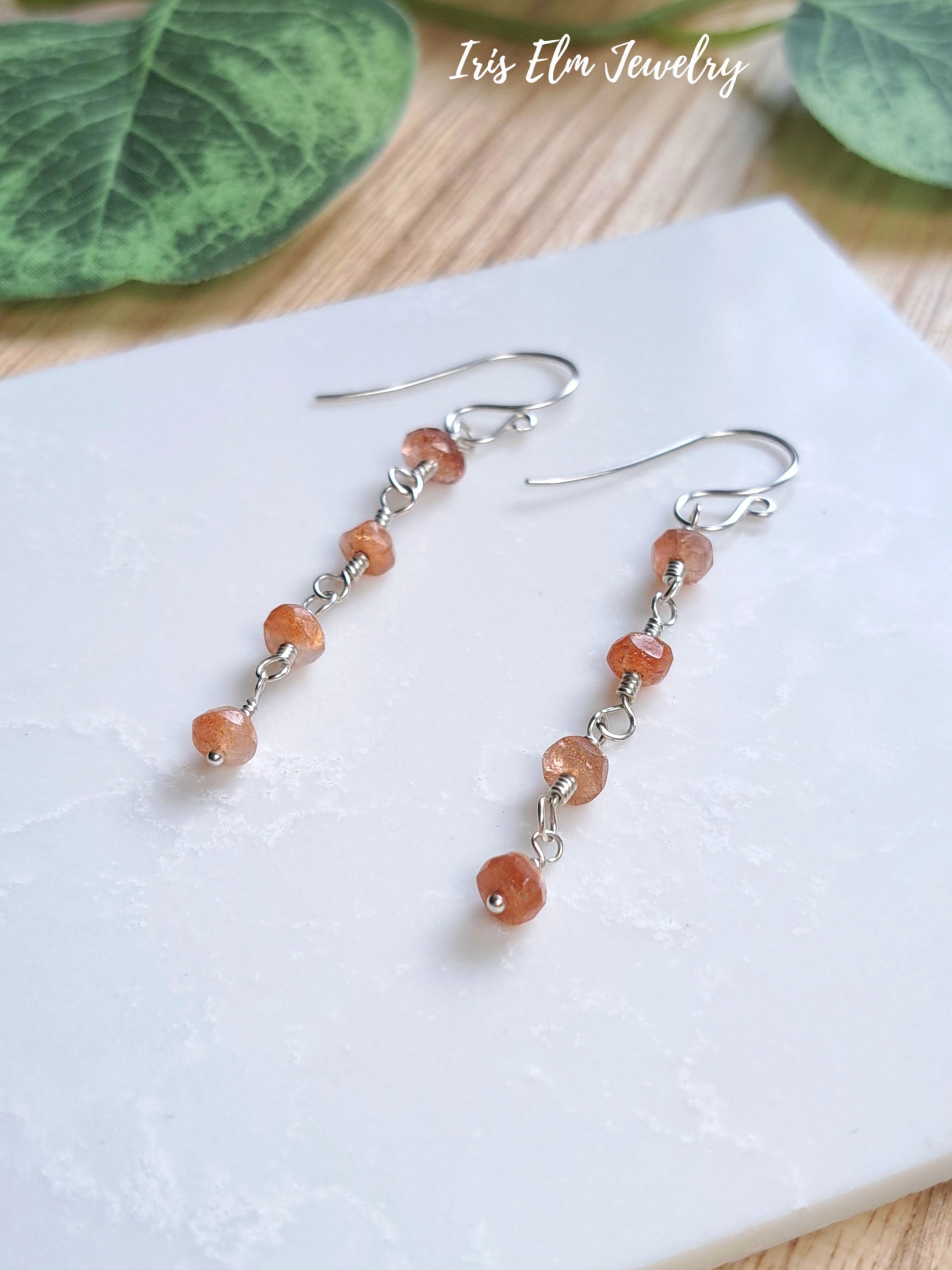 A pair of dangle earrings with small round orange-brown sunstone beads and silver hooks, displayed on a white textured surface with green leaves in the background. The text 'Iris Elm Jewelry' is visible in the upper right.