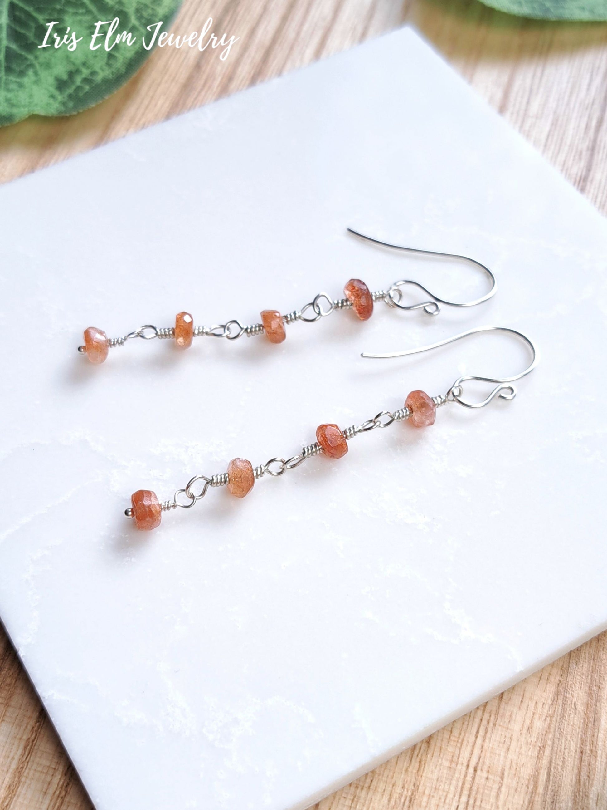 A pair of dangle earrings with small peach-orange translucent sunstone beads on silver wire hooks, displayed on a white square tile with a light wood surface and green leaves in the background. The text 'Iris Elm Jewelry' is visible in the top left corner.