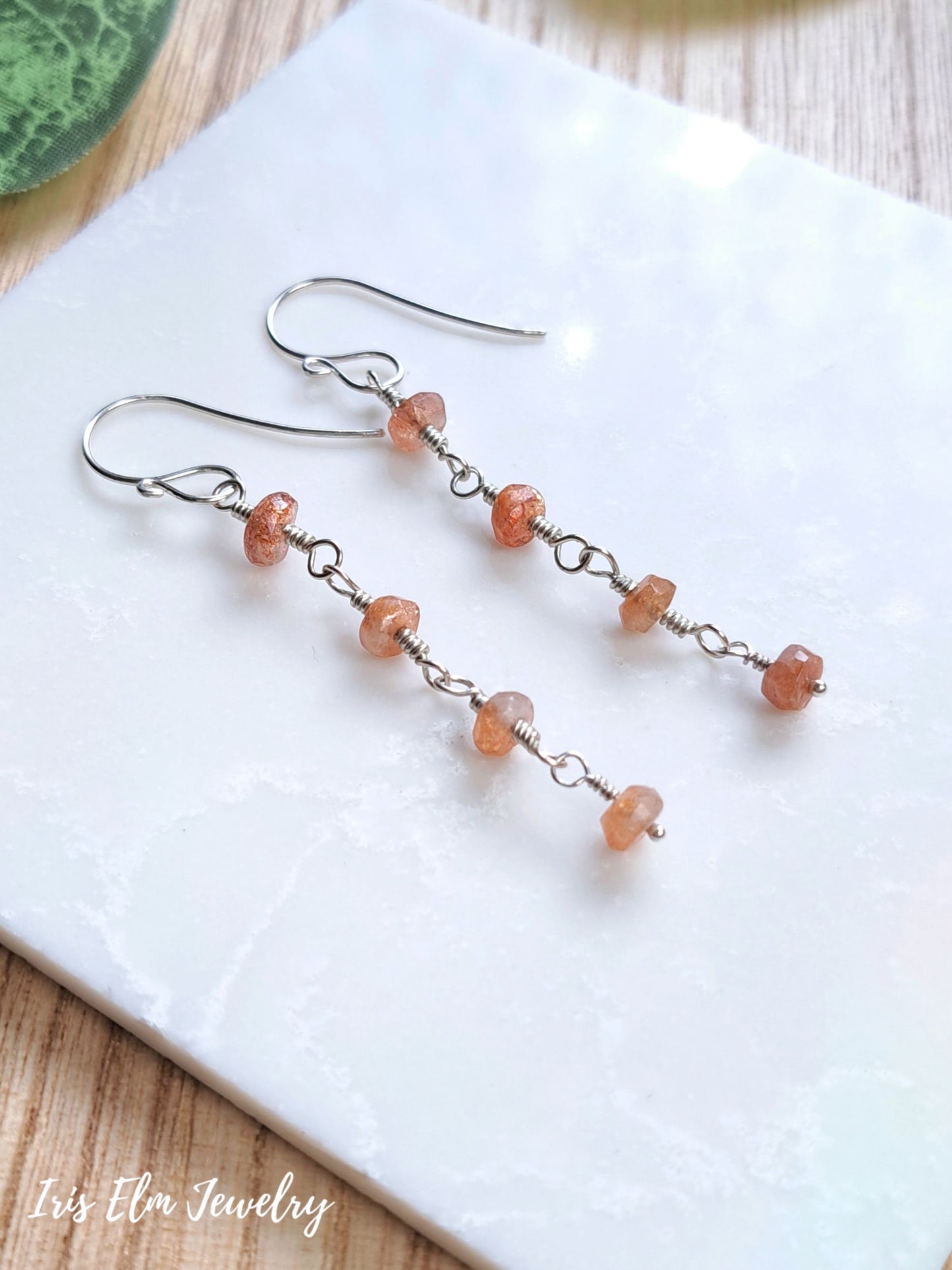 A pair of handmade dangle earrings with small peach-orange faceted sunstone beads and silver wire loops, displayed on a white textured tile with a wooden surface underneath. The brand name 'Iris Elm Jewelry' is visible in the bottom-left corner.