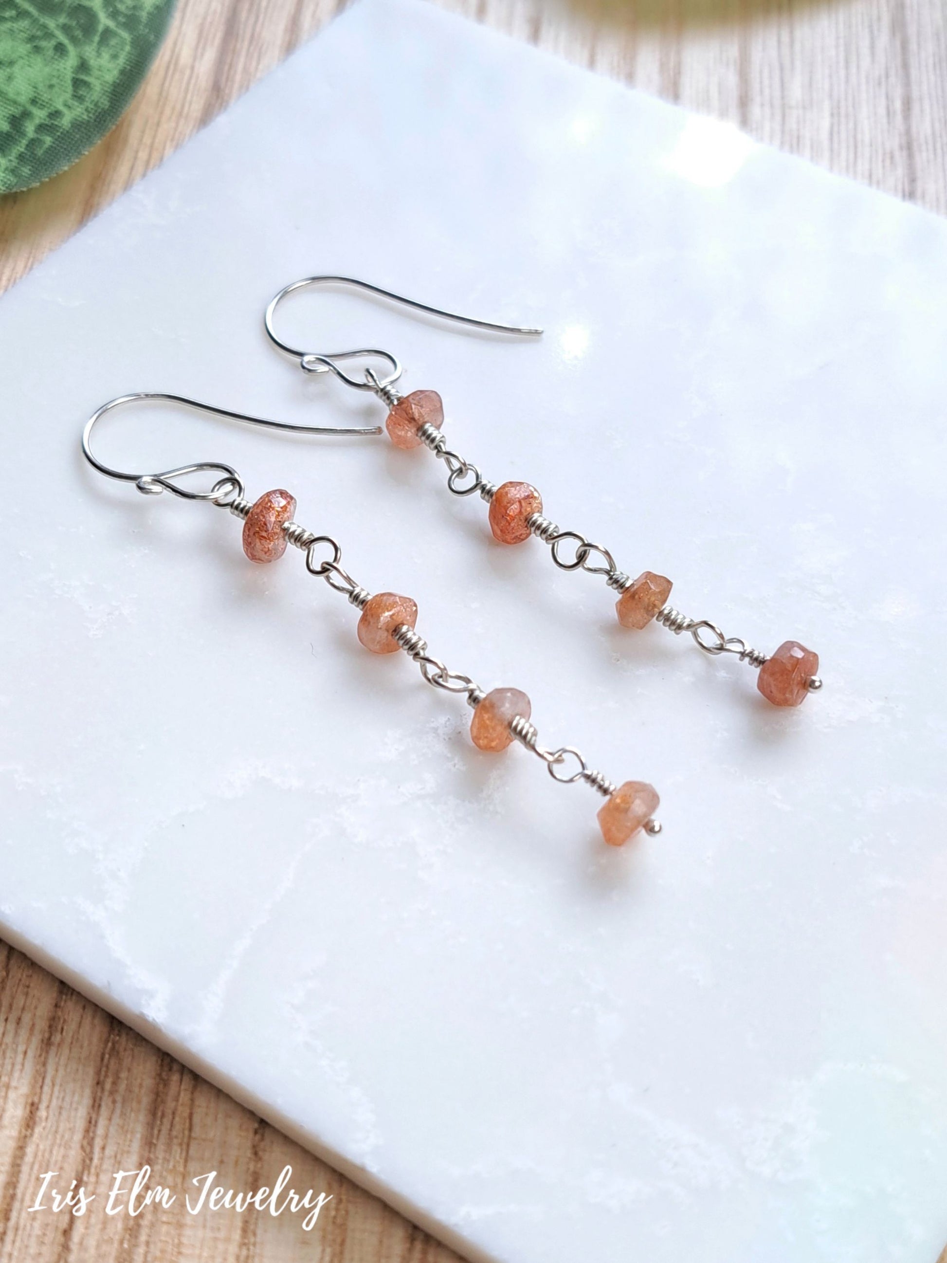 A pair of handmade dangle earrings with small peach-orange faceted sunstone beads and silver wire loops, displayed on a white textured tile with a wooden surface underneath. The brand name 'Iris Elm Jewelry' is visible in the bottom-left corner.