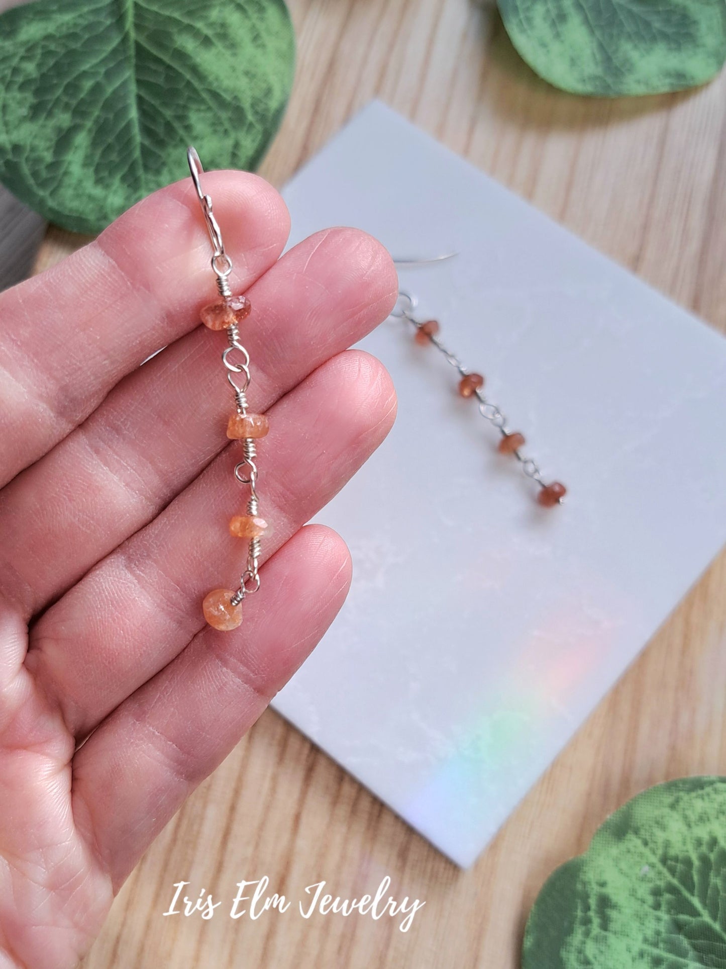 A close-up of a hand holding a single dangling beaded earring made of small peach-orange beads and silver wire loops, with a second matching earring resting on a white card on a wooden surface. Green leaves are partially visible around the scene, and the text 'Iris Elm Jewelry' appears at the bottom.