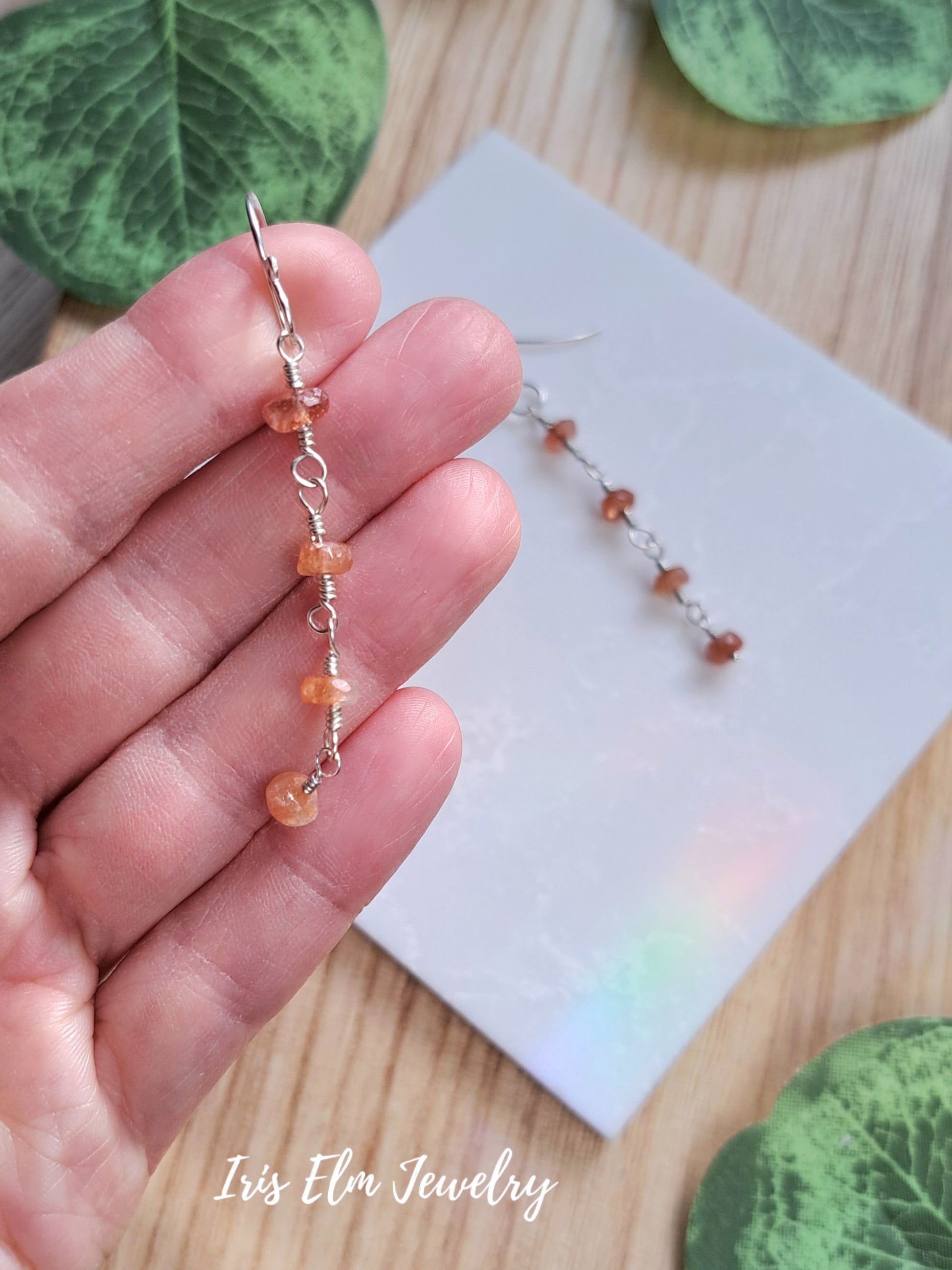 A close-up of a hand holding a single dangling beaded earring made of small peach-orange beads and silver wire loops, with a second matching earring resting on a white card on a wooden surface. Green leaves are partially visible around the scene, and the text 'Iris Elm Jewelry' appears at the bottom.