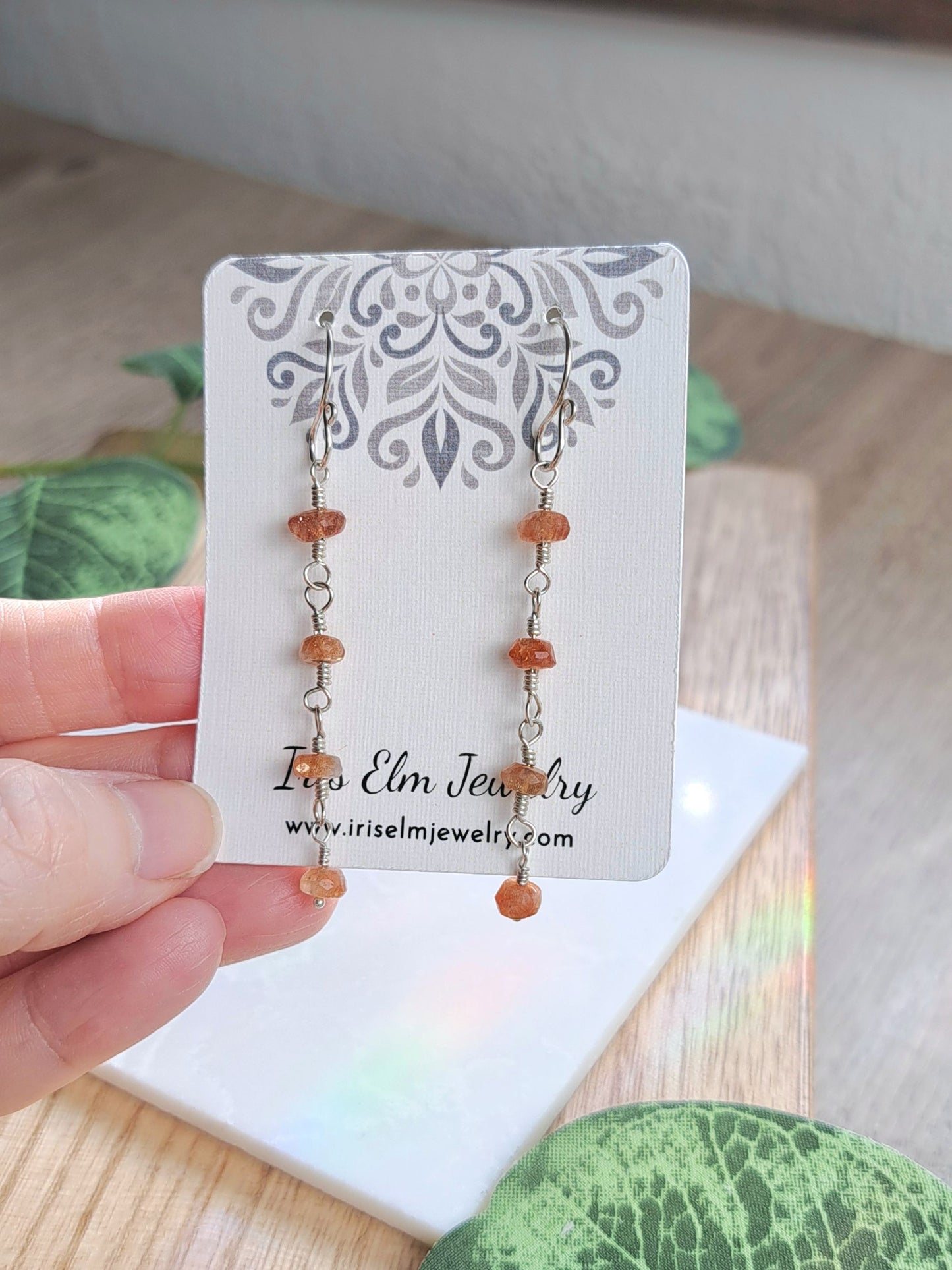 A hand holds a white jewelry display card featuring a pair of dangling earrings with small amber-orange beads and silver-toned links. The card shows the brand name Iris Elm Jewelry.